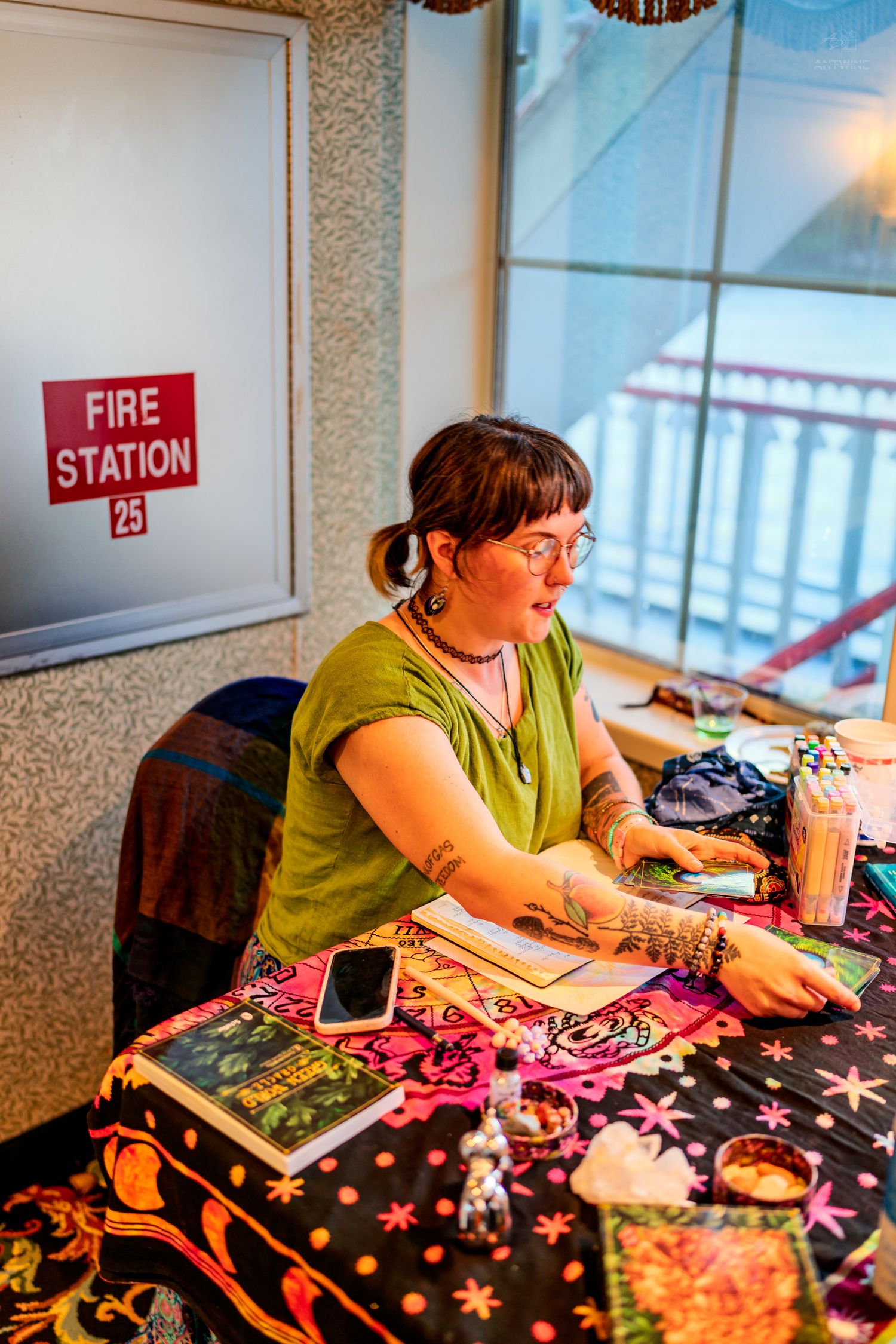 Woman with tattoos reads tarot cards at a decorated table near a Fire Station 25 sign at a psychic fair.
