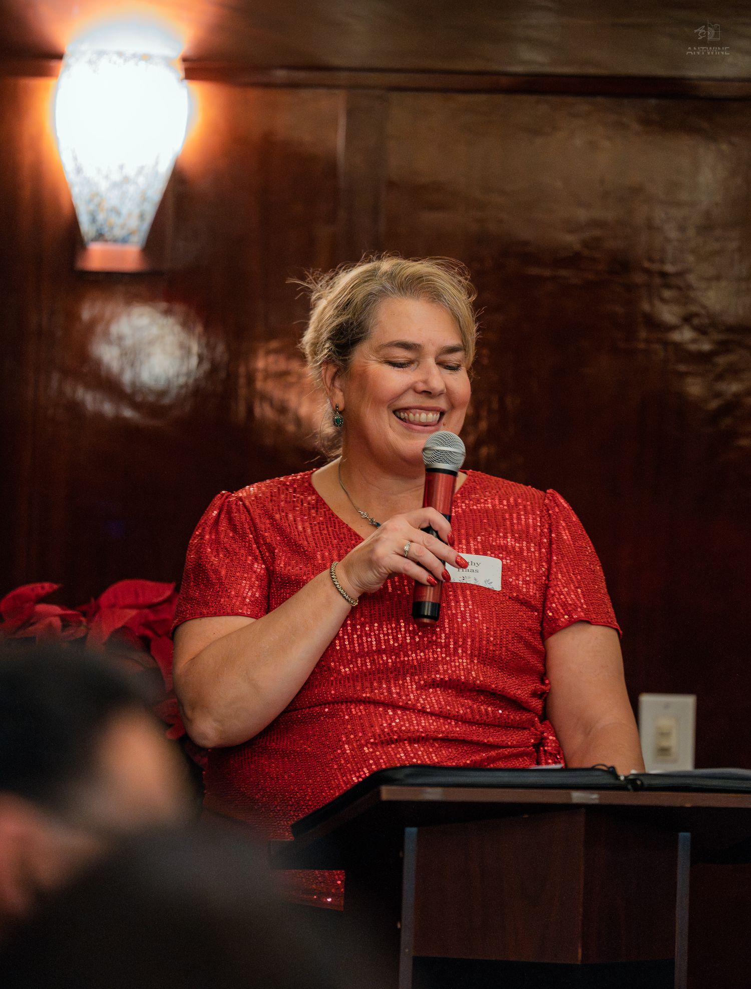 Woman in red sequin dress laughing while speaking into microphone at indoor event.