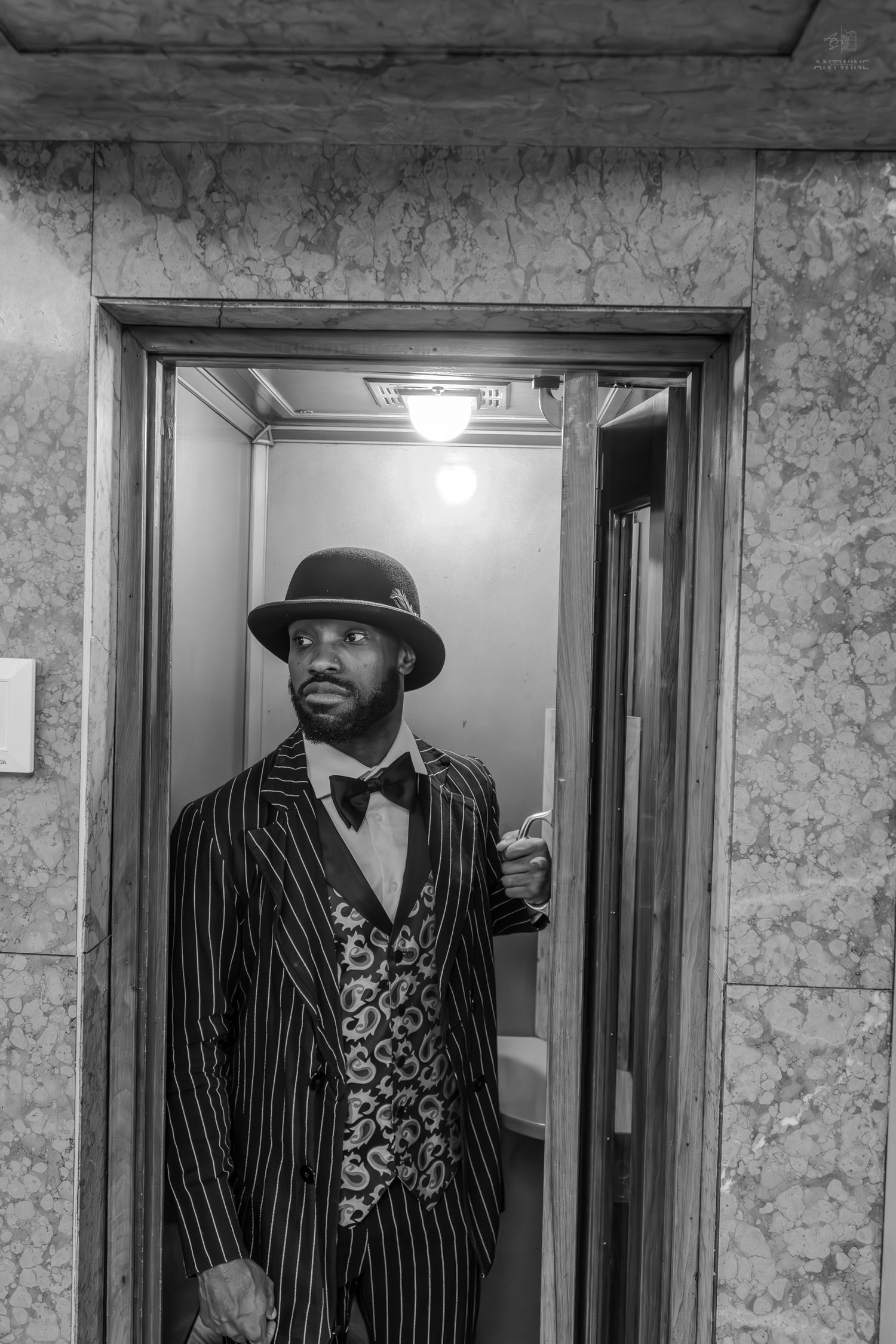 Man in pinstripe suit, bow tie, and fedora hat standing in an elevator doorway, black and white photo.