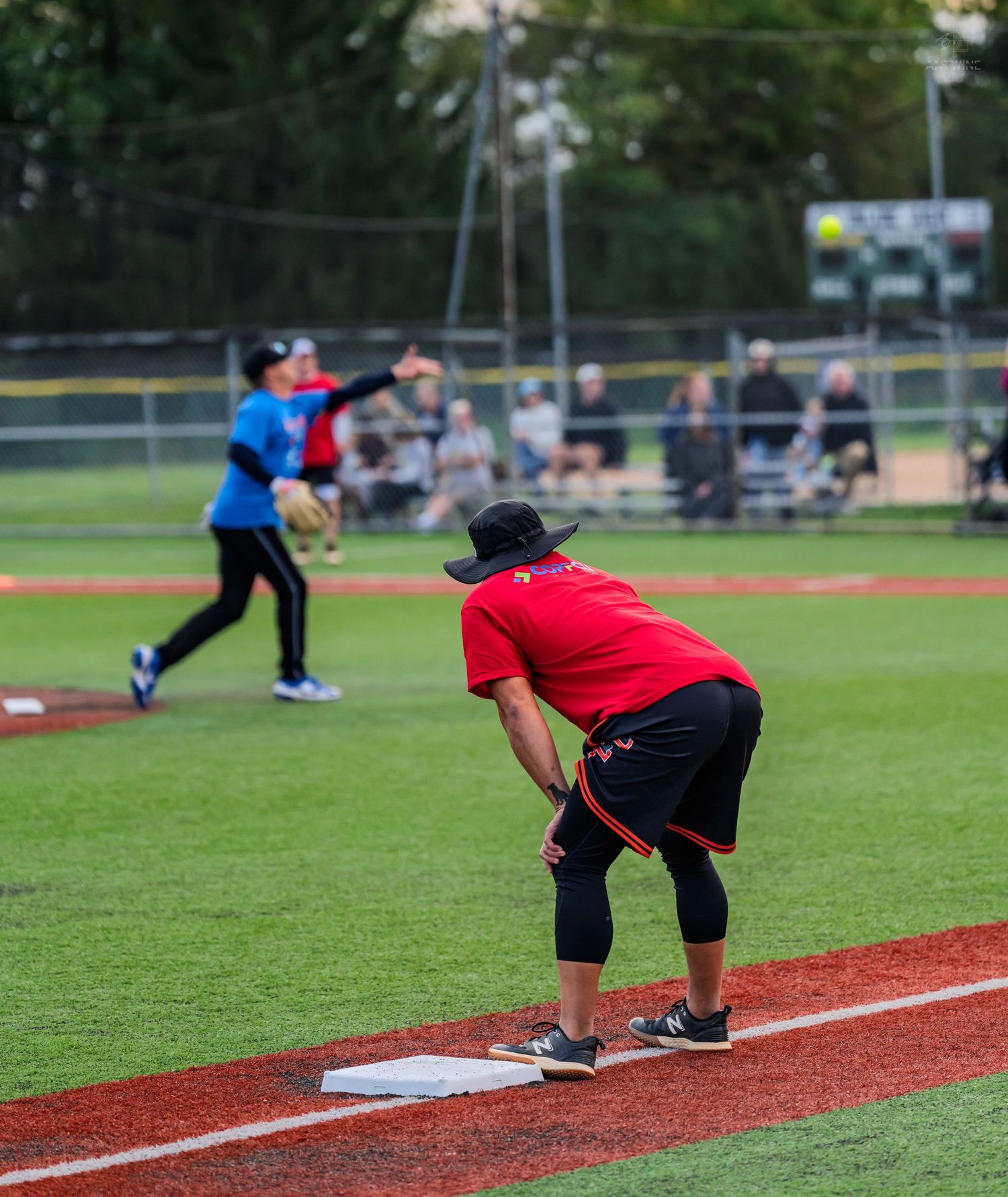 Female softball player in red shirt crouching at first base while pitcher throws ball to batter.