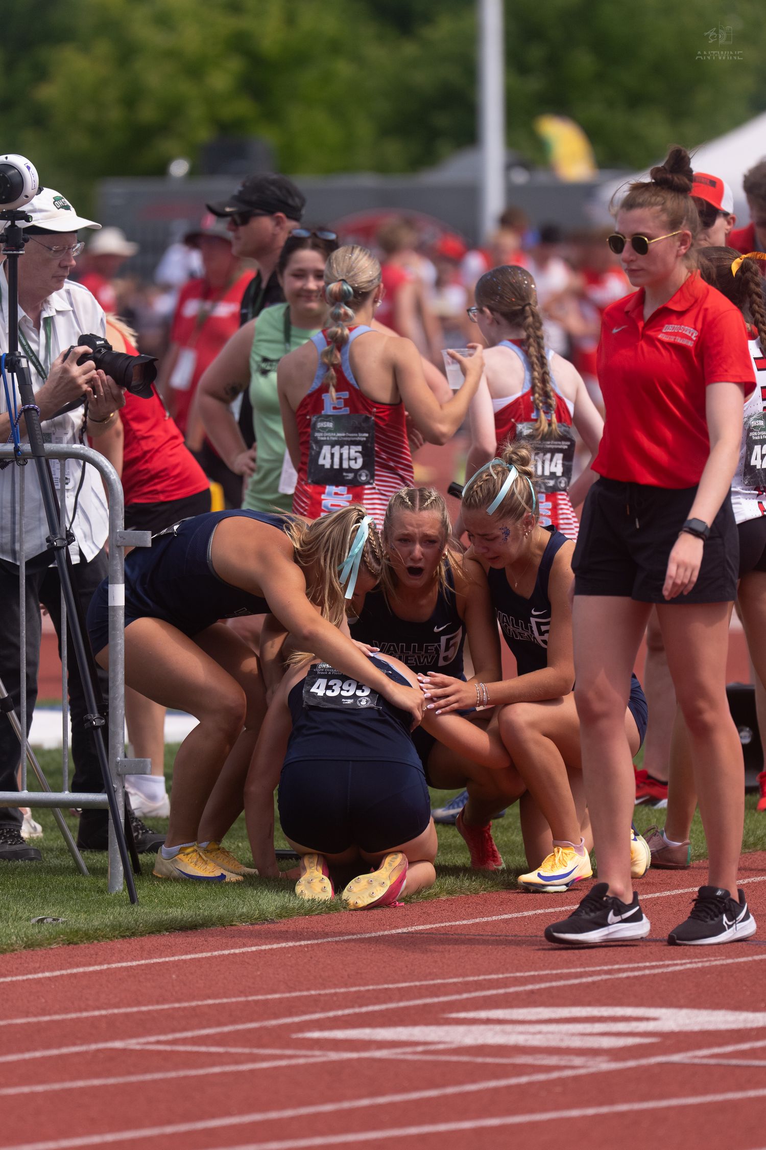 Emotional female athlete wearing bib 4393 is consoled by teammates while kneeling on the track post-race.