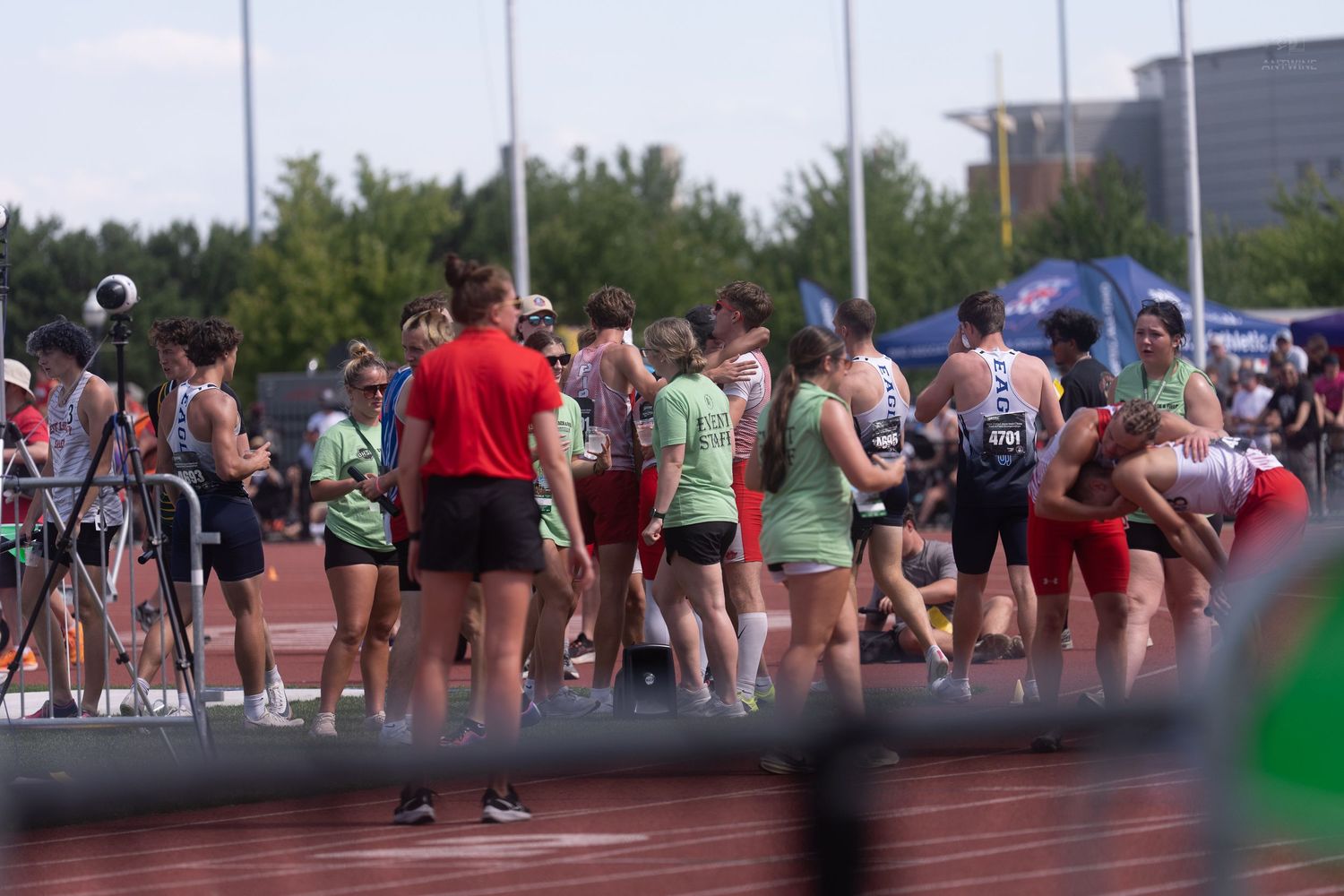 Large group of track and field athletes in colorful uniforms gathered on a red track during a meet.