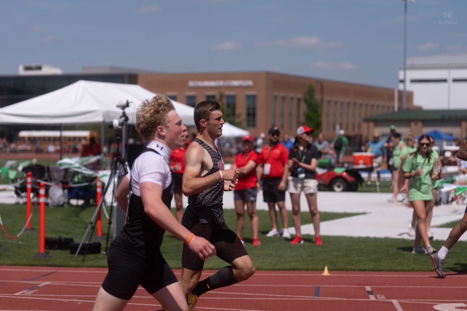 Two male track athletes run side by side on a red outdoor track during a field competition.