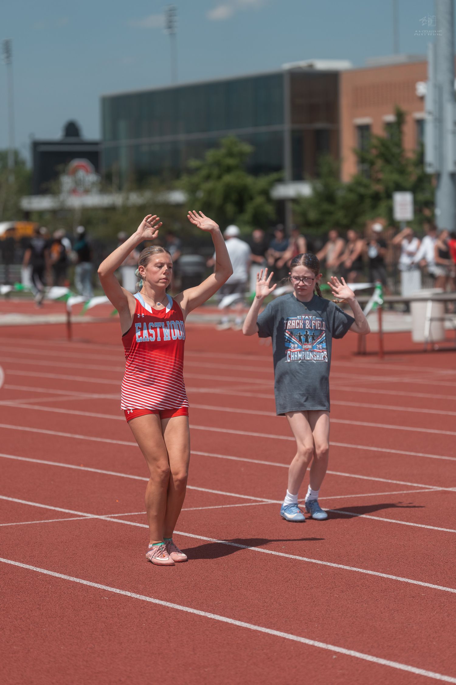 Two female athletes celebrate with raised hands on red track at sunny outdoor track and field competition.