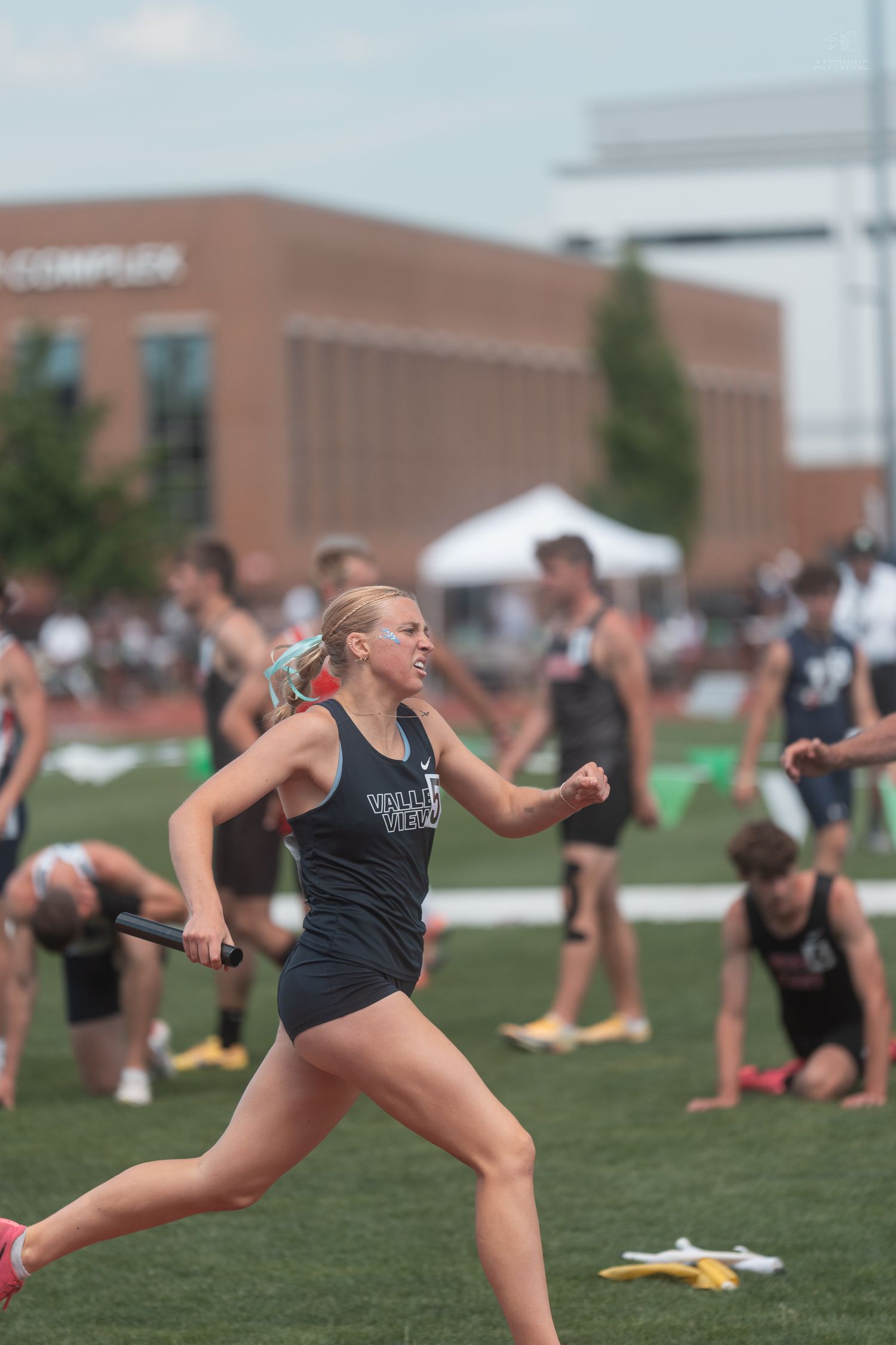 Blonde female track athlete runs with a relay baton at an outdoor high school track and field competition.