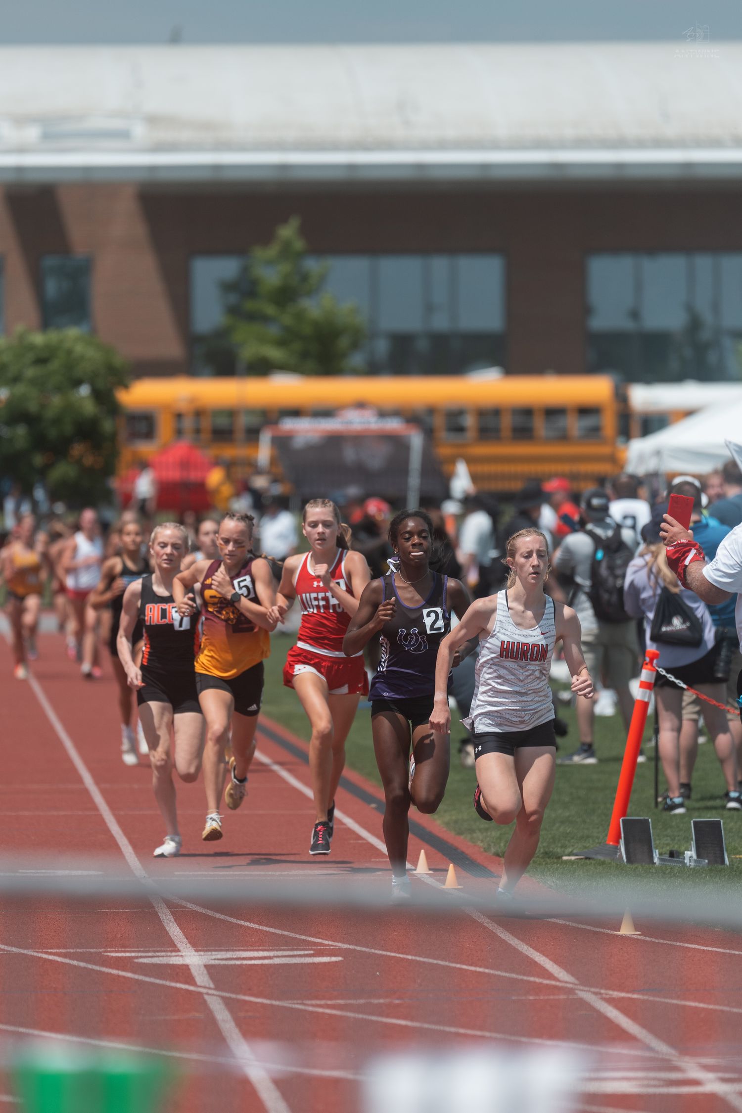 Large group of female runners compete in a middle distance track race at an outdoor high school championship meet.