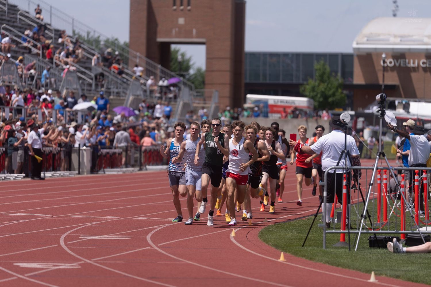 Runners approach the finish line at a high school track meet with a large crowd cheering in the stadium.