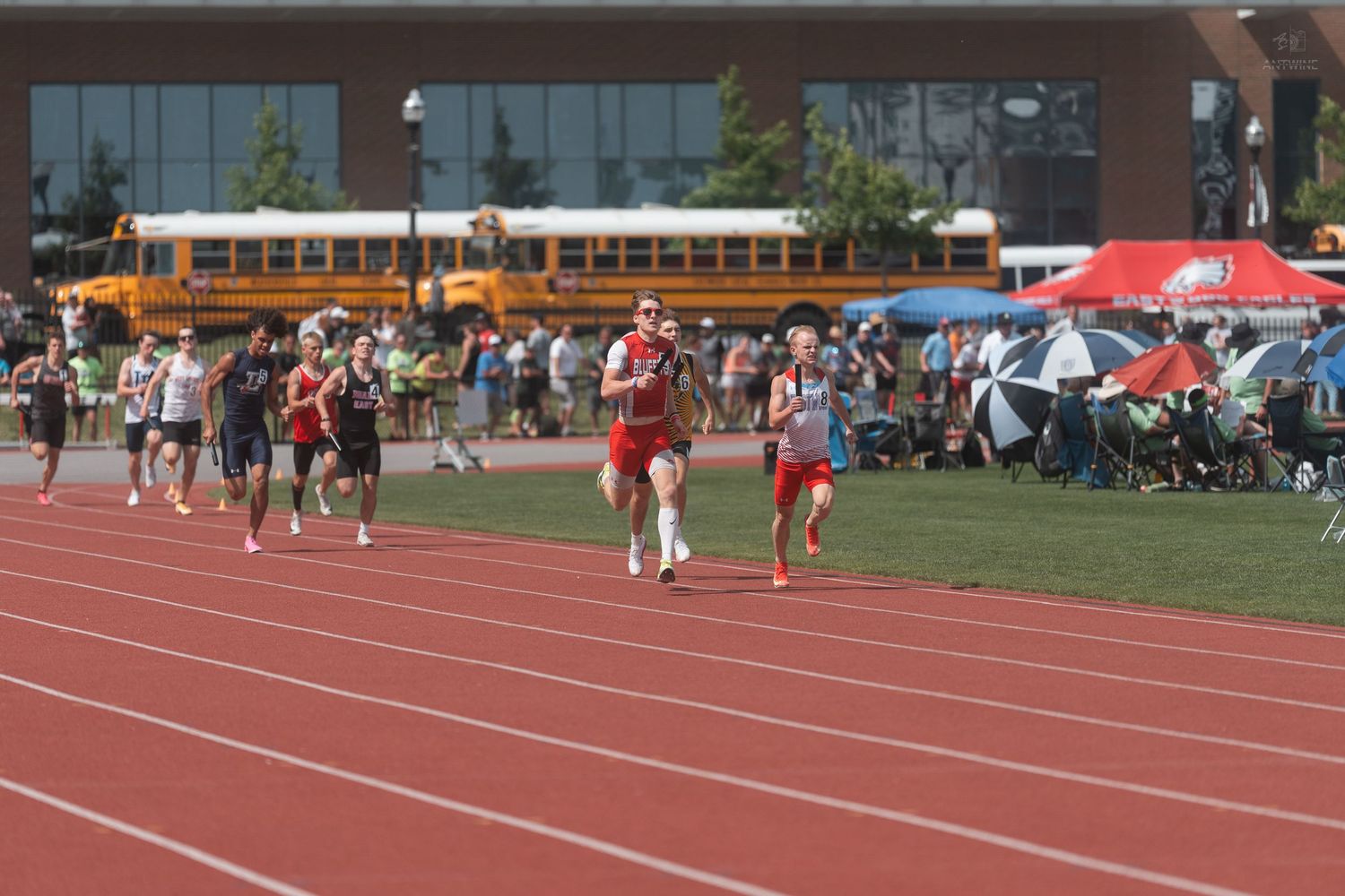 High school runners race on a red track with a yellow school bus and crowd visible in the background.