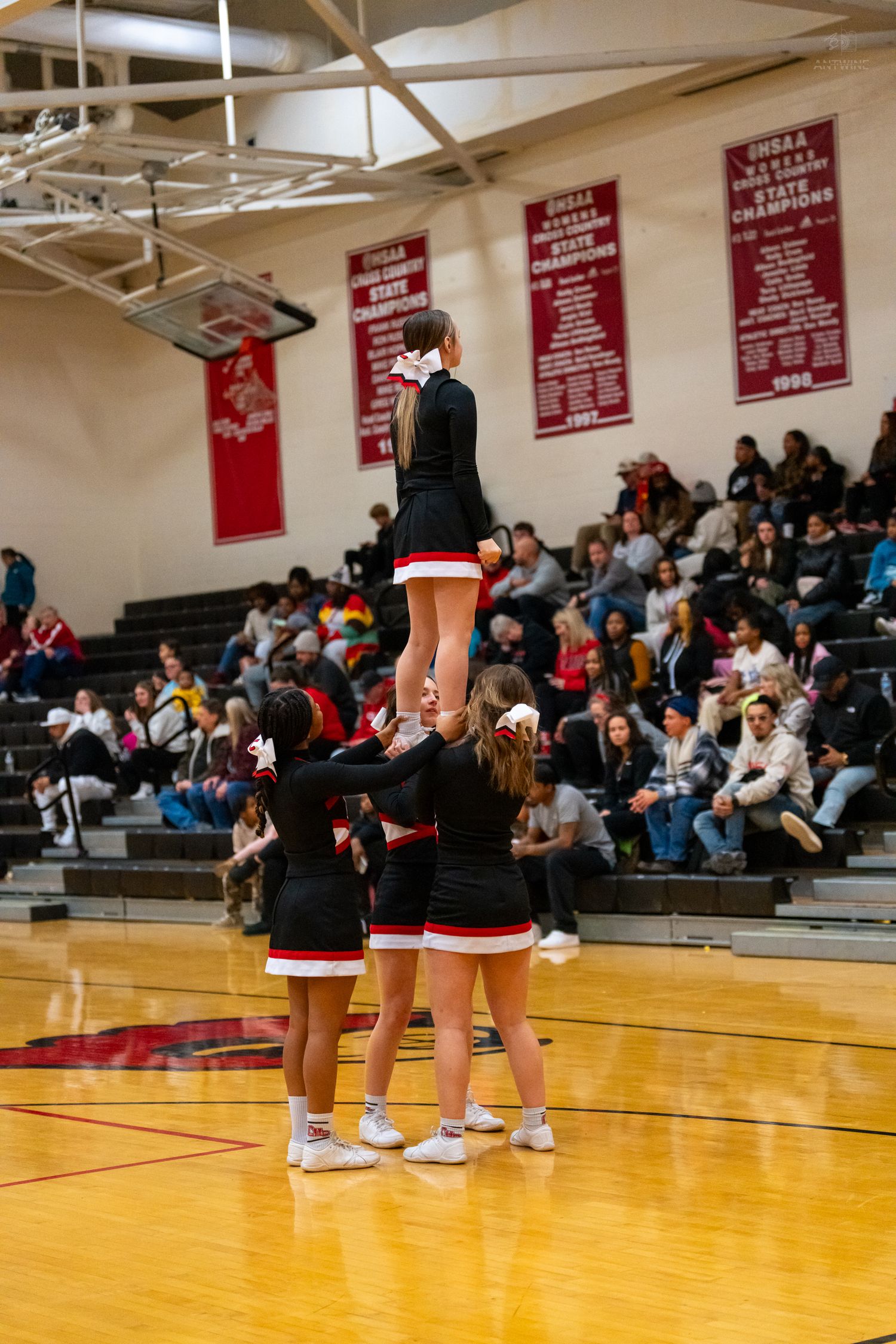 High school cheerleaders perform a pyramid stunt in front of a packed gymnasium during a basketball game.