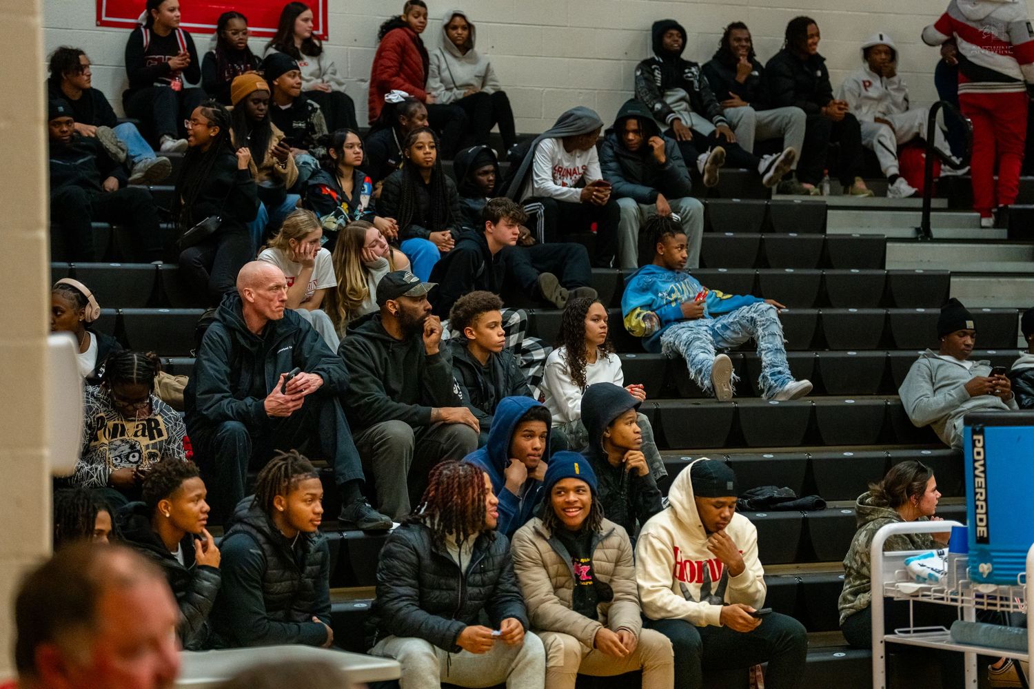 Diverse crowd of fans seated in gymnasium bleachers watching a high school basketball game.