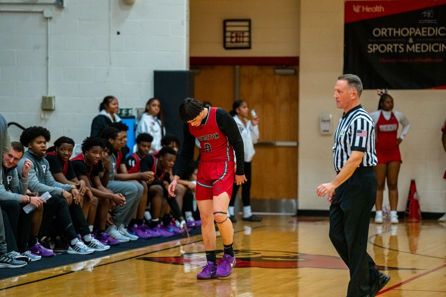 A basketball player in a red uniform bends over near a referee during a high school basketball game.