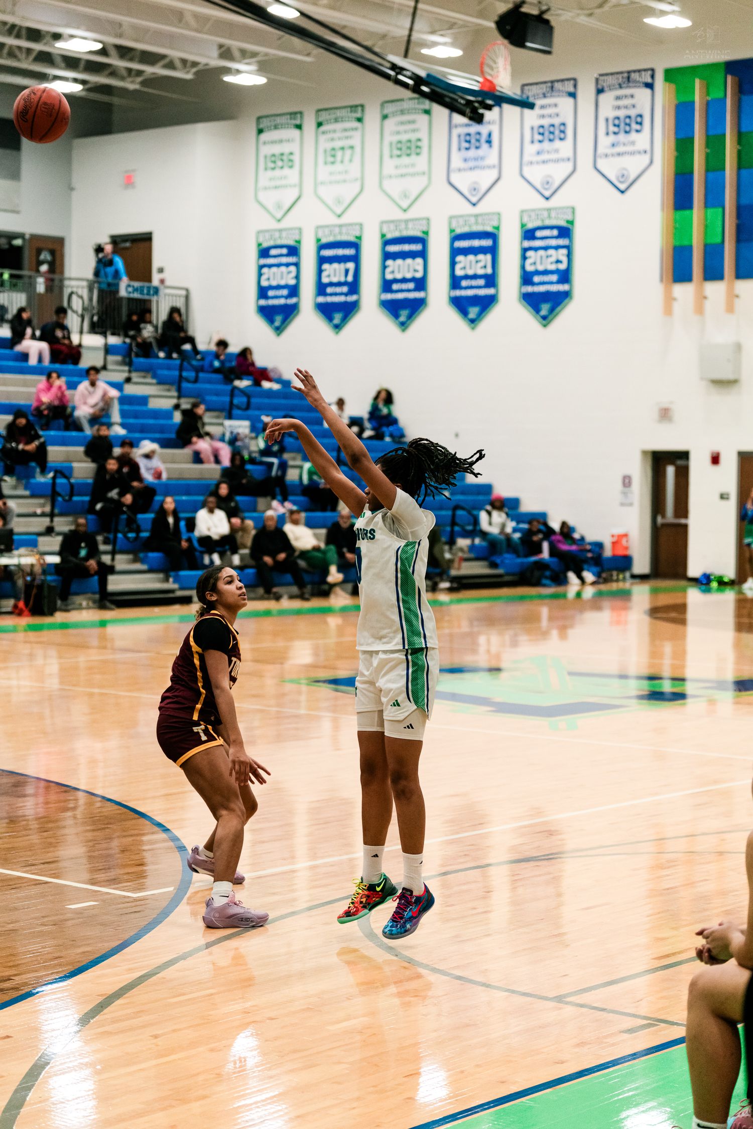 Female basketball player in green Warriors uniform shoots a jump shot over a maroon-uniformed defender in high school game.