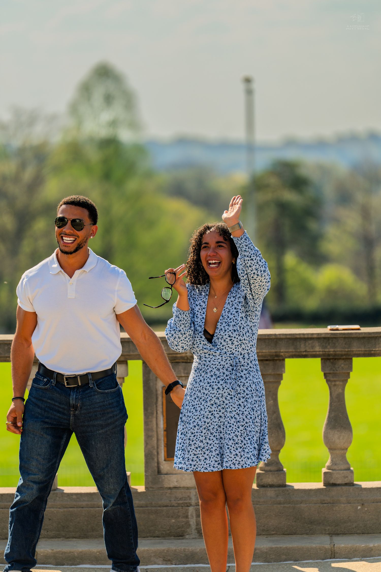 A laughing couple poses on a sunny stone terrace, woman waving with green park landscape behind.