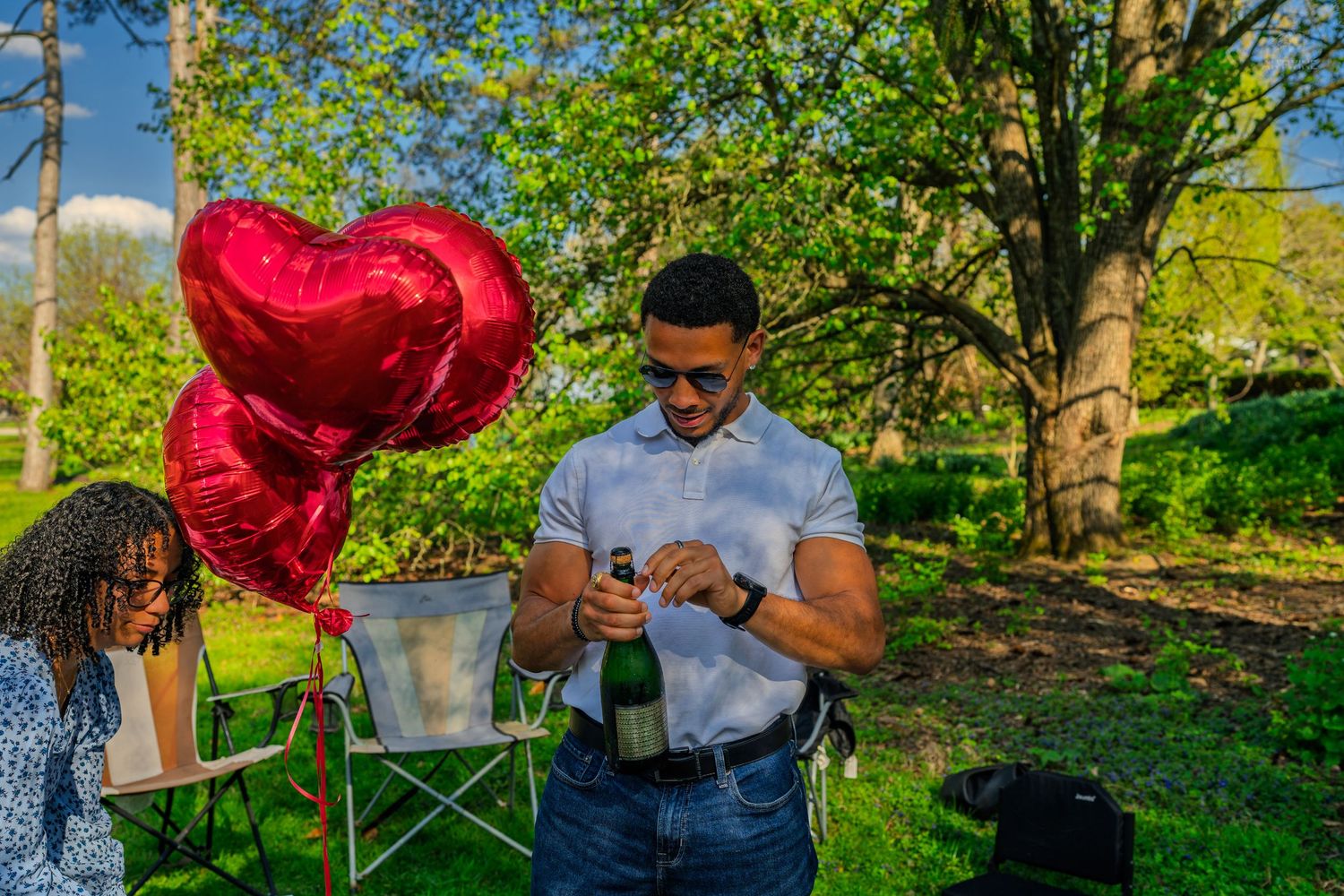 Man opening a champagne bottle outdoors near red heart balloons and a woman seated in a park setting.