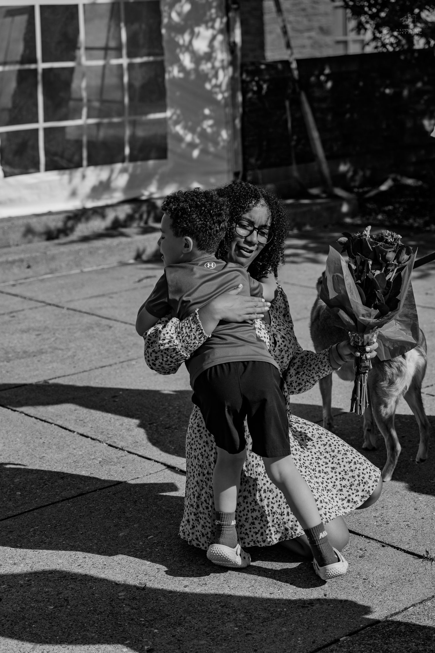 A laughing woman in a floral dress embraces a young child outdoors in black and white.
