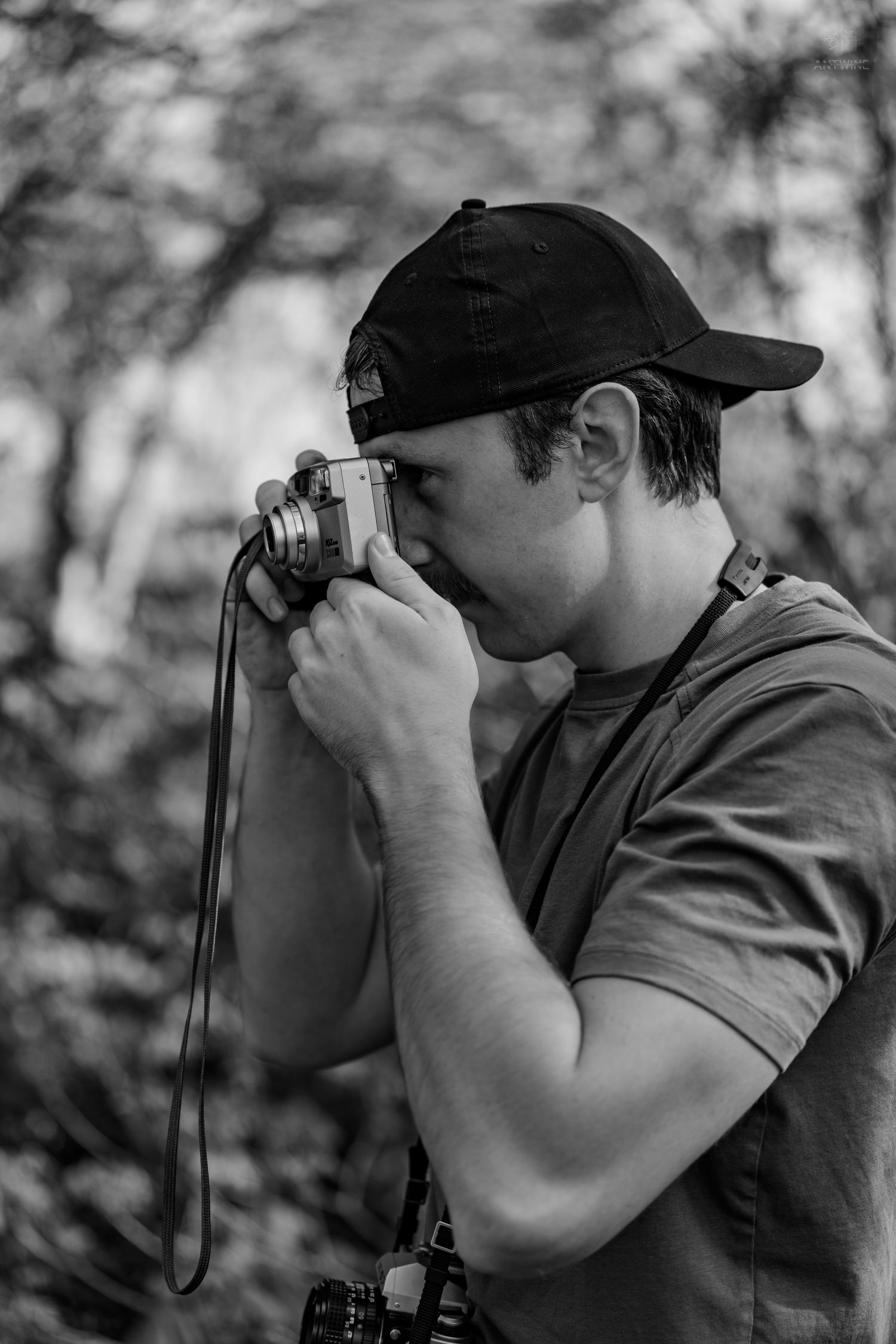 Black and white photo of young man in cap shooting with vintage film camera in forest.