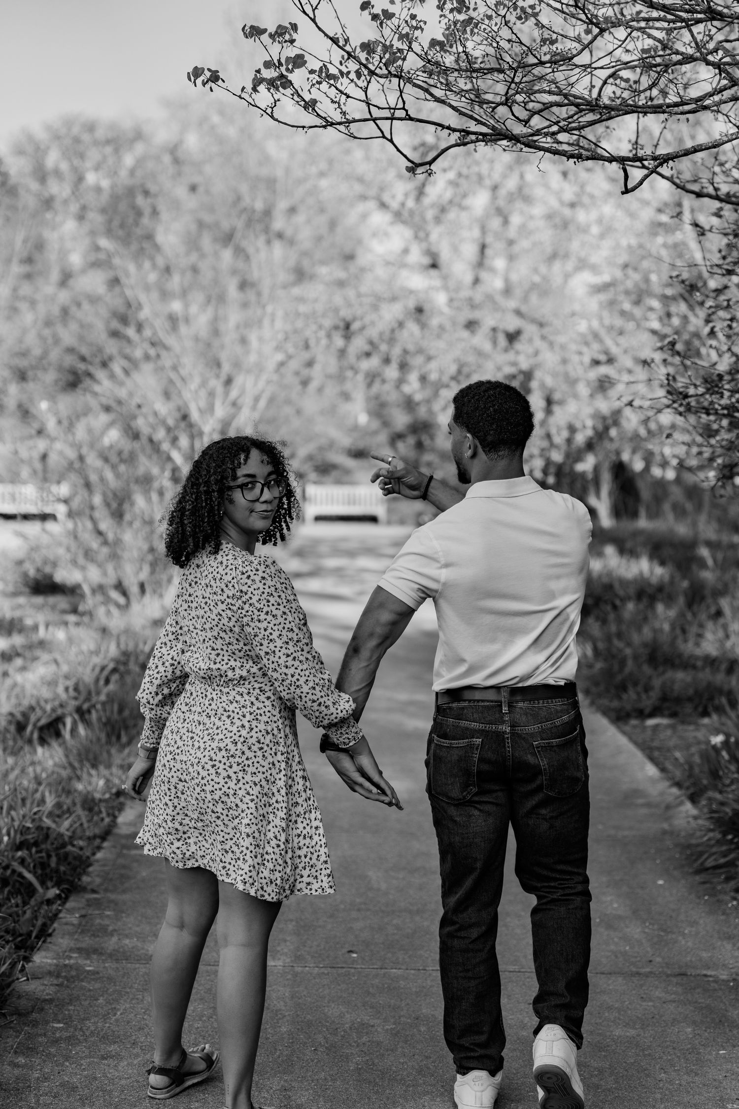 Black and white photo of a couple walking hand in hand on a tree-lined park pathway.