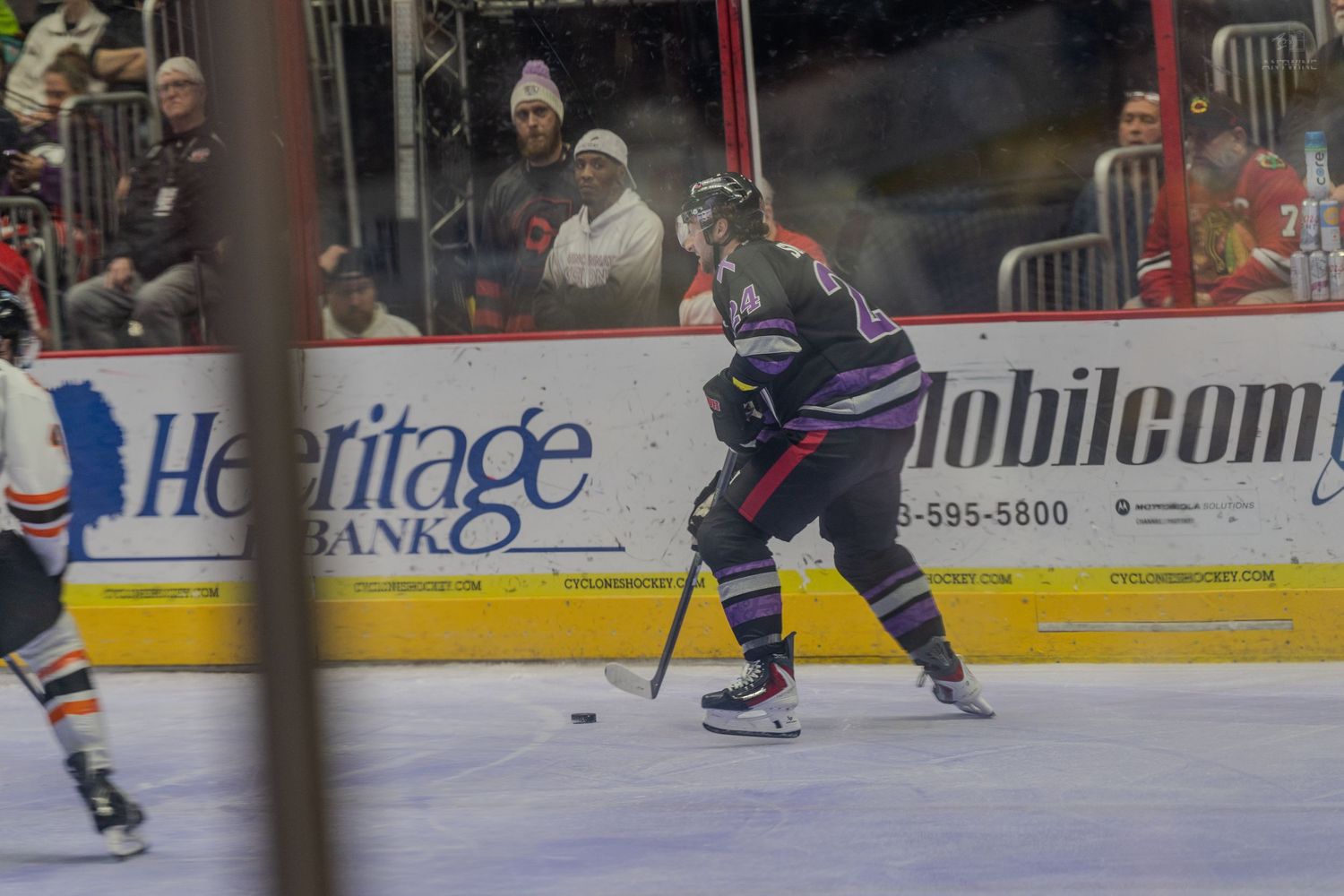 A hockey player in purple skates with the puck along the boards during a minor league game.
