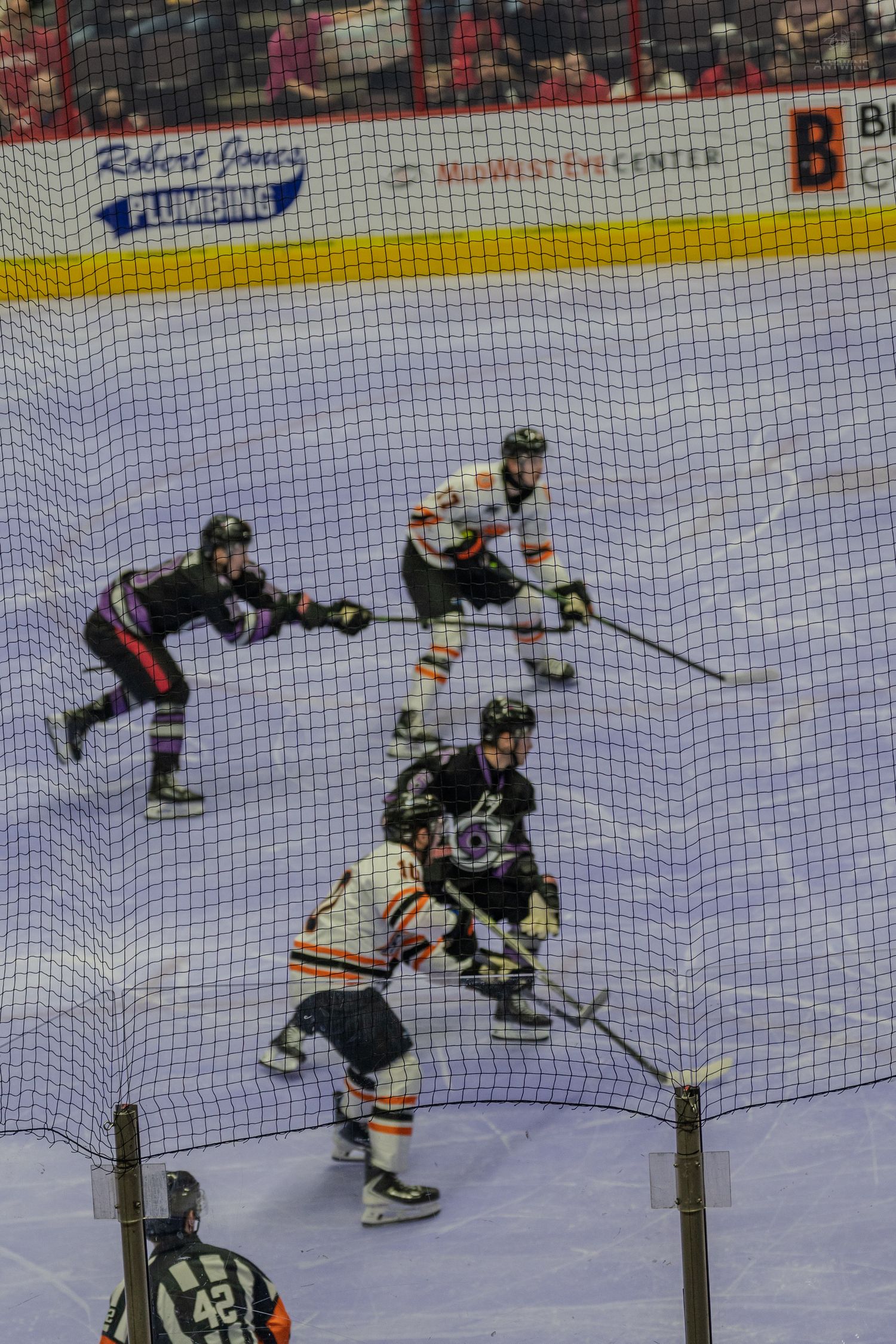 Four ice hockey players compete for the puck near the boards as a referee watches the play.