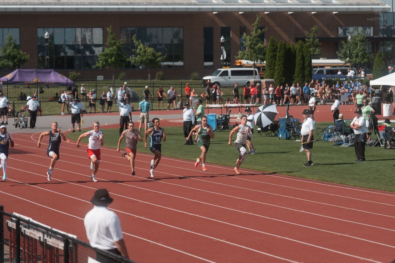 Male athletes sprint down a red track during an outdoor track and field competition on a clear sunny day.