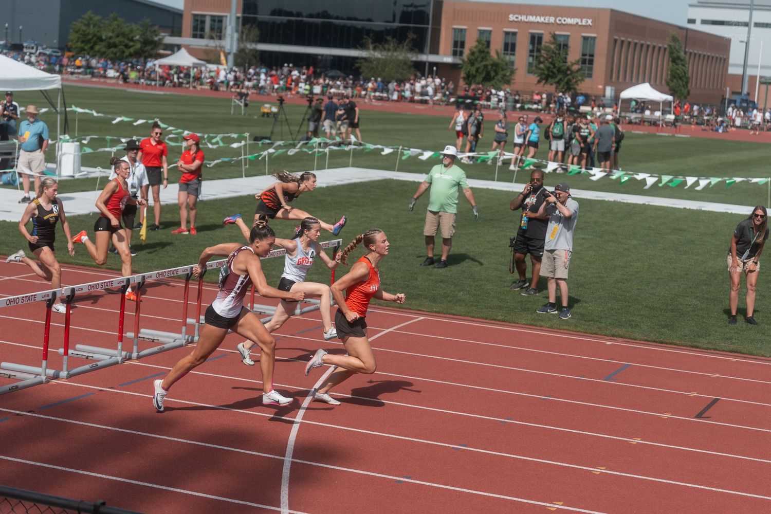 Female athletes sprint toward finish line in hurdles race at outdoor track and field championship event.