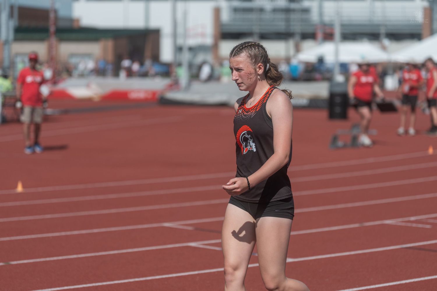 Female athlete in black and orange uniform runs on a red track at a competitive track meet.