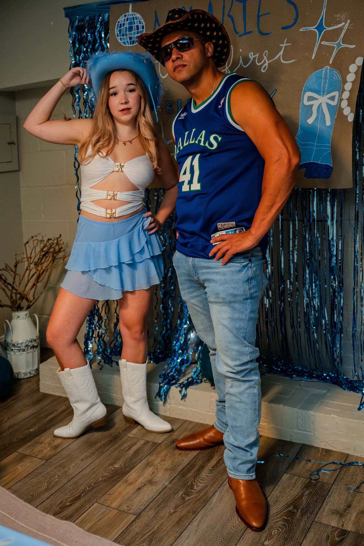 Duo in Western and Dallas Mavericks themed outfits pose against blue streamers at birthday party.