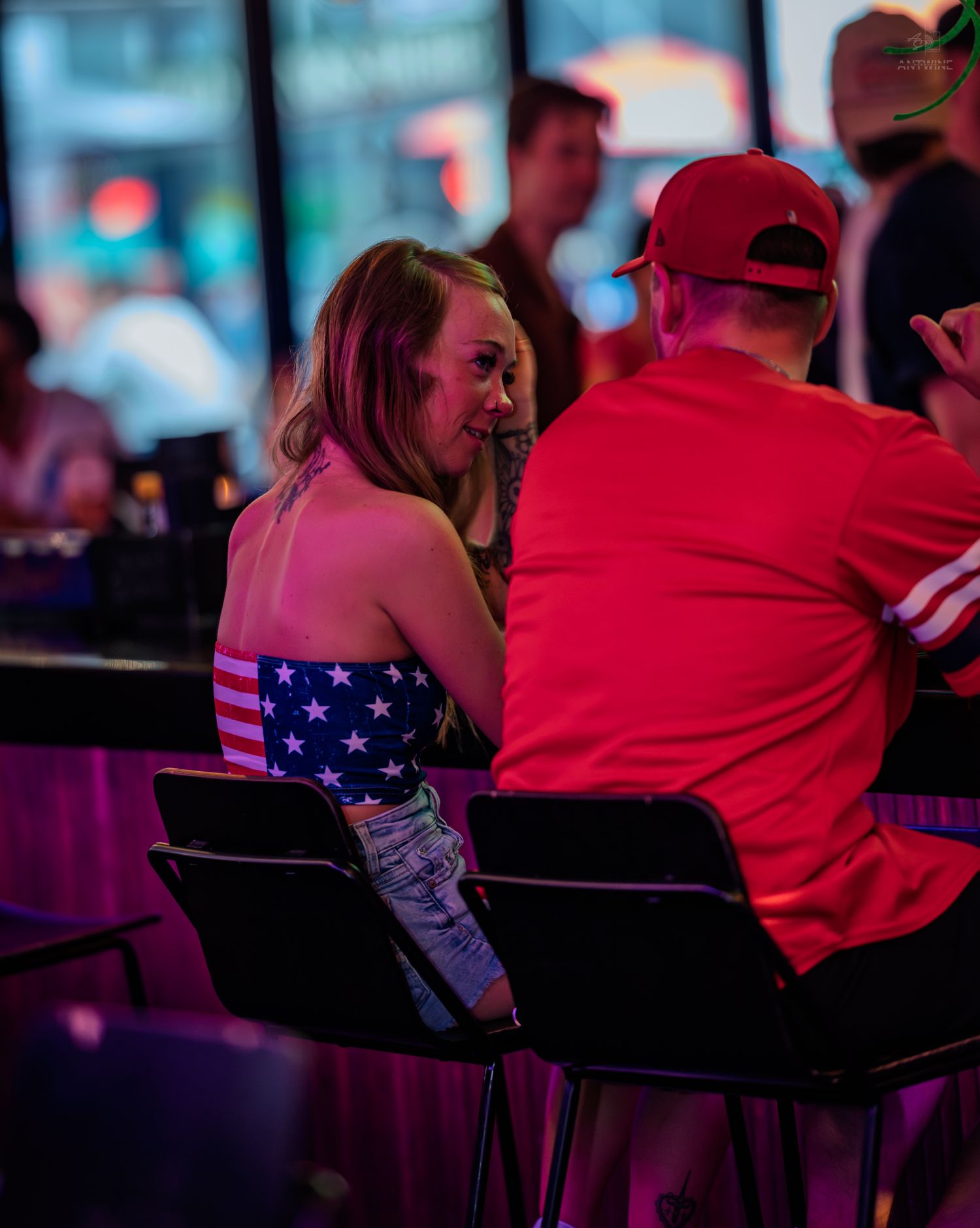 A woman in an American flag top sits with a man in red at a nightclub bar.