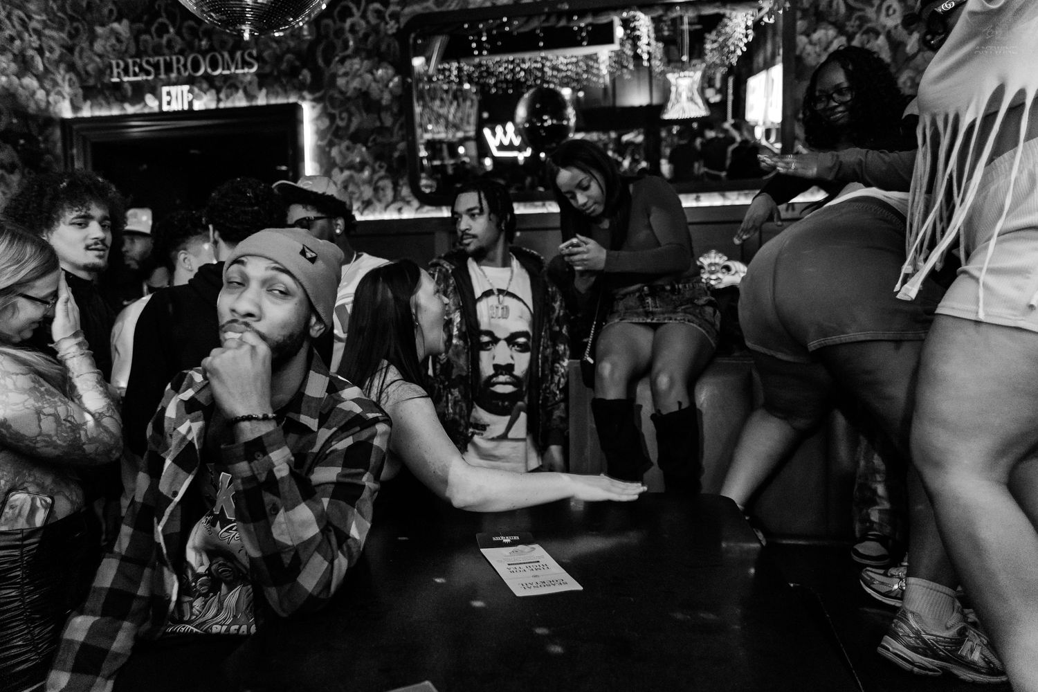 Diverse group of people gathered at a bar counter with restroom sign visible in black and white photo.