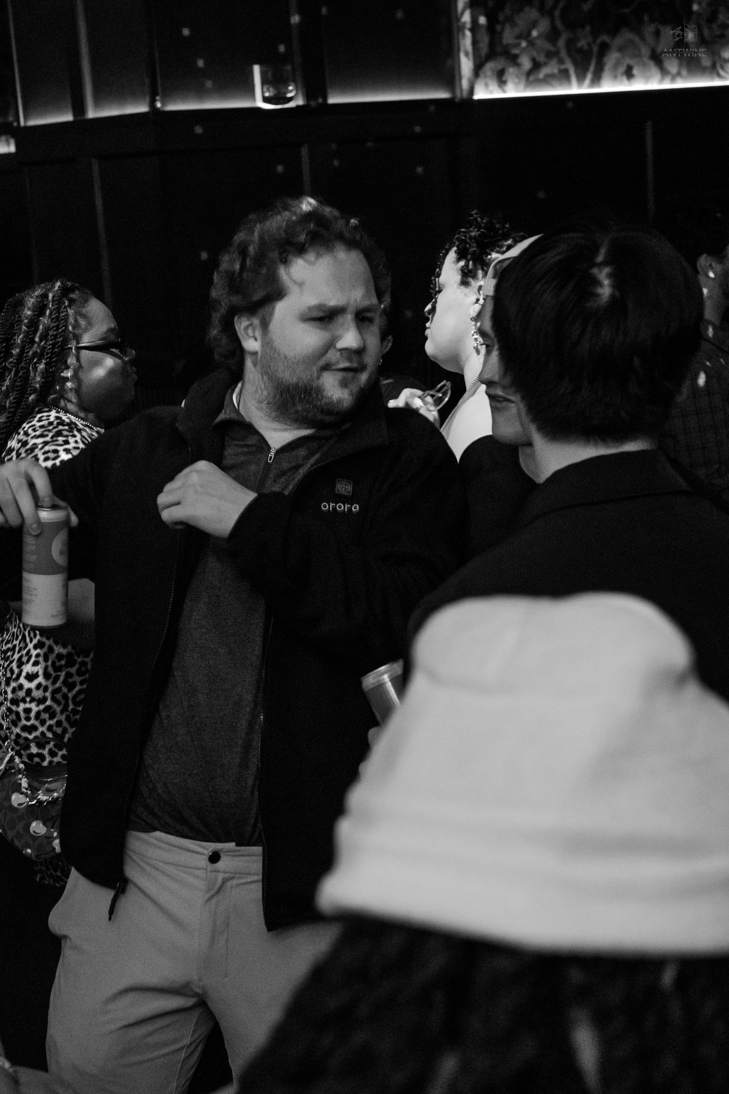 A bearded man in a zip-up jacket socializes with others on a dimly lit nightclub dance floor.