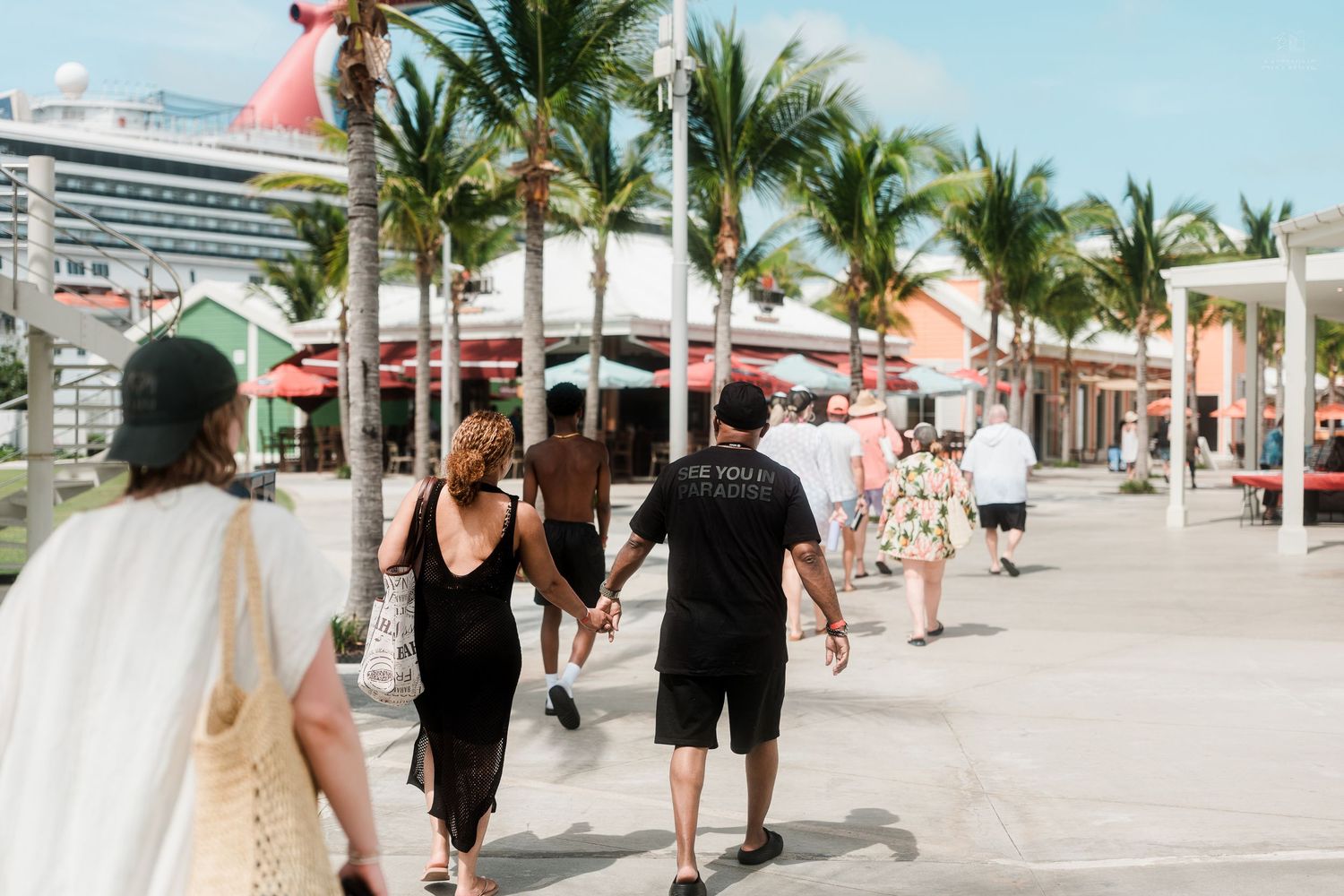 Tourists walk along a palm-lined promenade near a cruise ship port in a tropical destination.