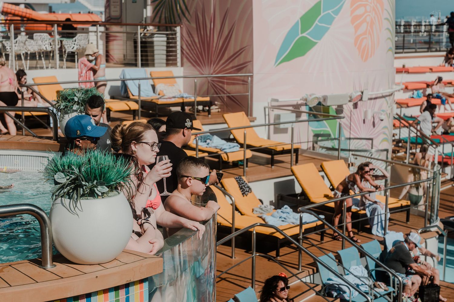 Couple relaxing by cruise ship pool deck surrounded by lounge chairs and tropical themed decor.