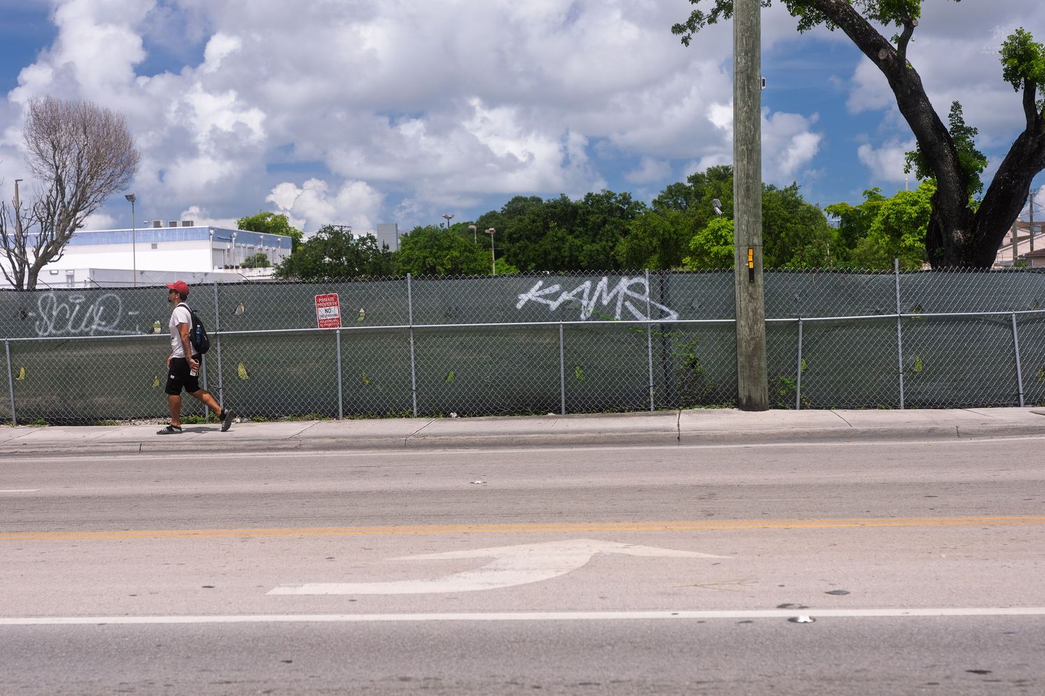 Person walking past a chain-link fence covered in graffiti tags along a urban street sidewalk.