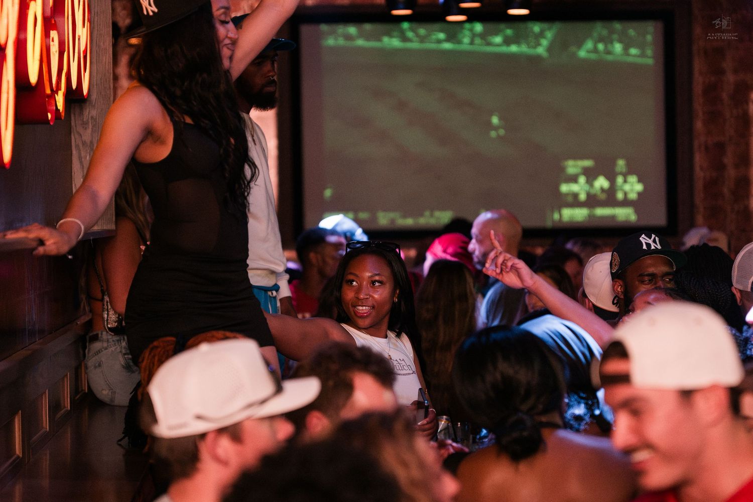 Crowded nightclub party with people dancing and socializing under large projection screens.