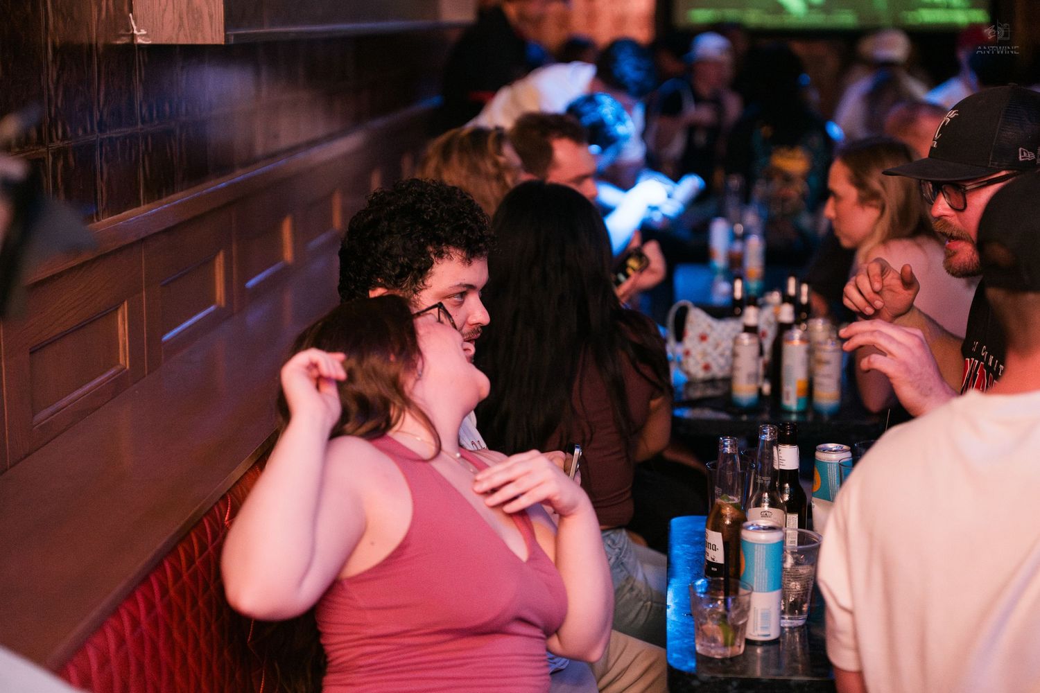 Woman in pink top laughing and socializing at a dimly lit bar with bottles and drinks on the counter.