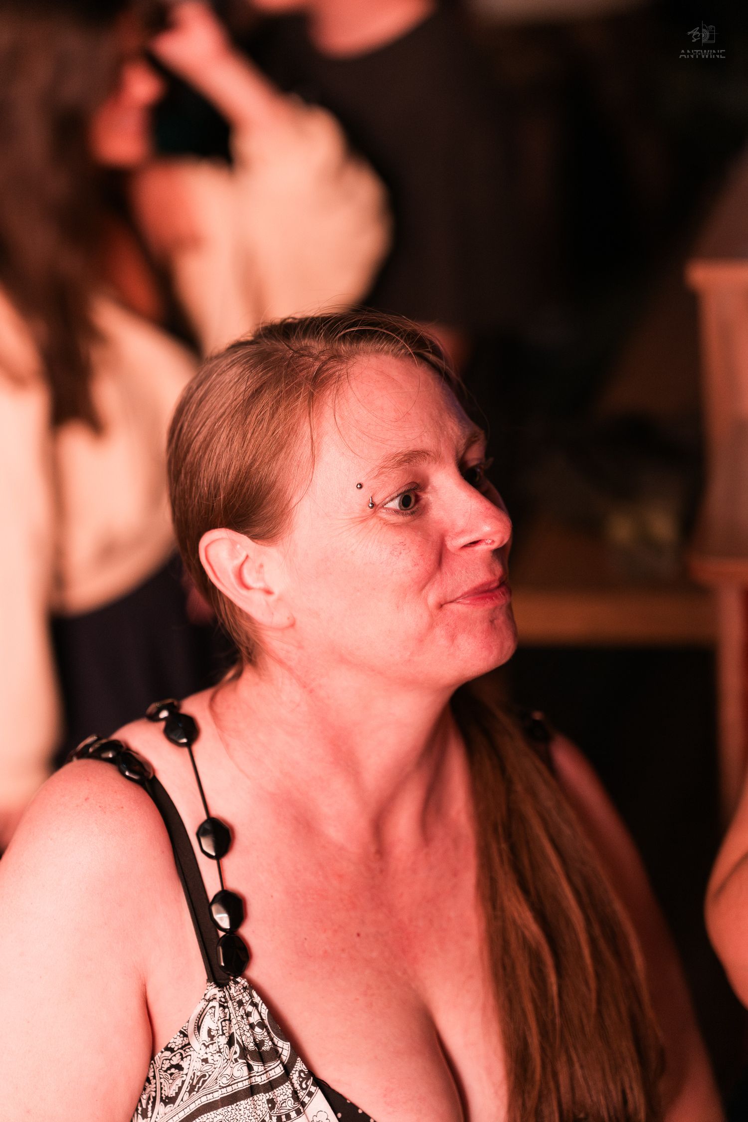 A middle-aged woman with auburn hair wearing a black beaded necklace gazes upward at a dimly lit social event.