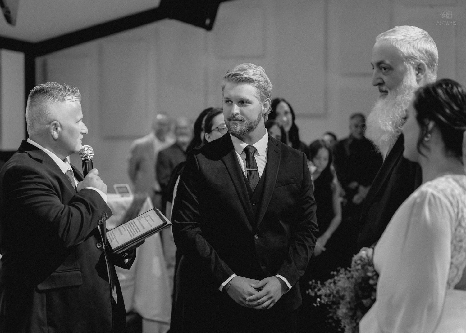 Groom in black suit exchanges vows with officiant and white-bearded minister at elegant indoor wedding ceremony.