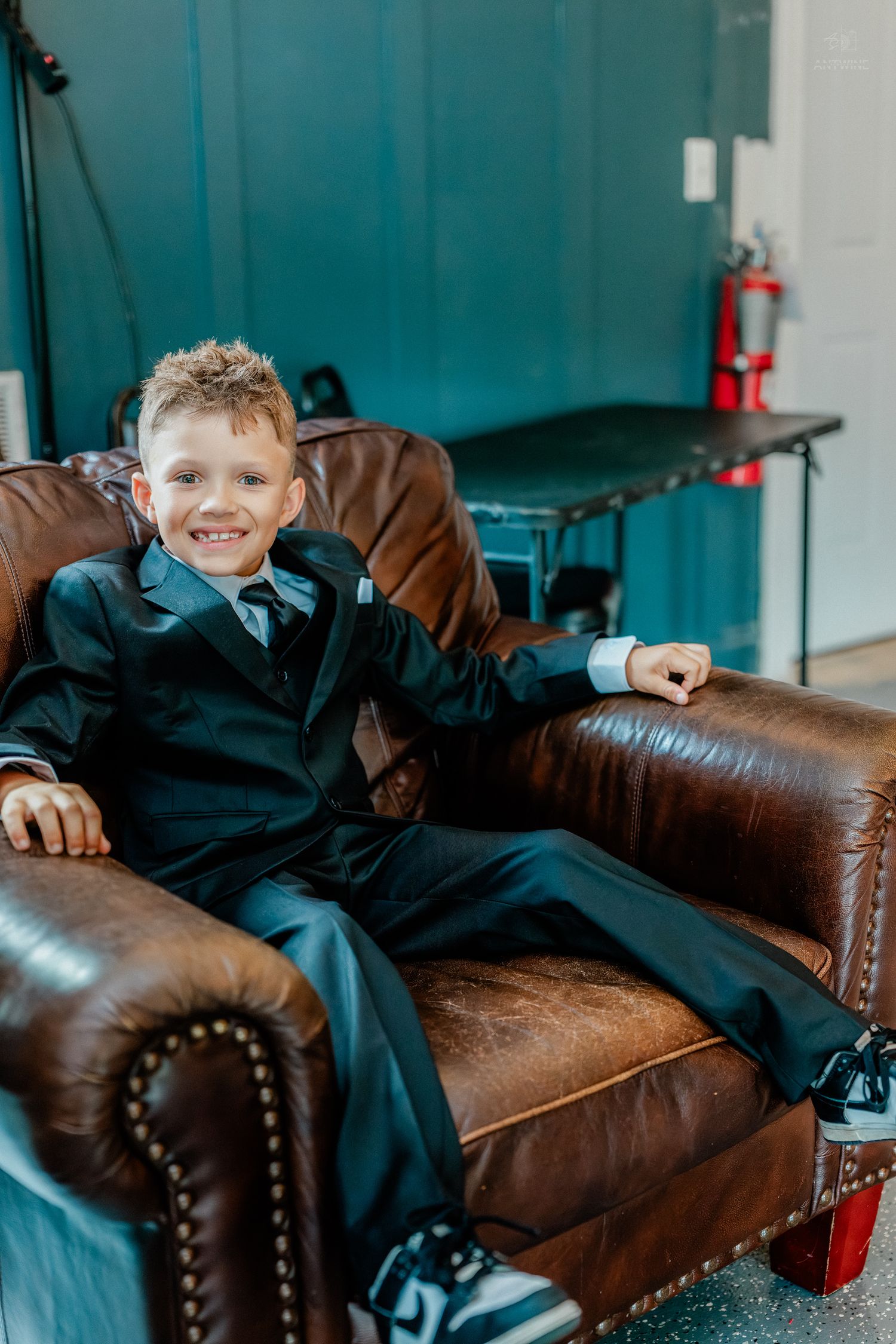 A smiling boy in a black suit relaxes confidently in a brown leather armchair at a wedding.
