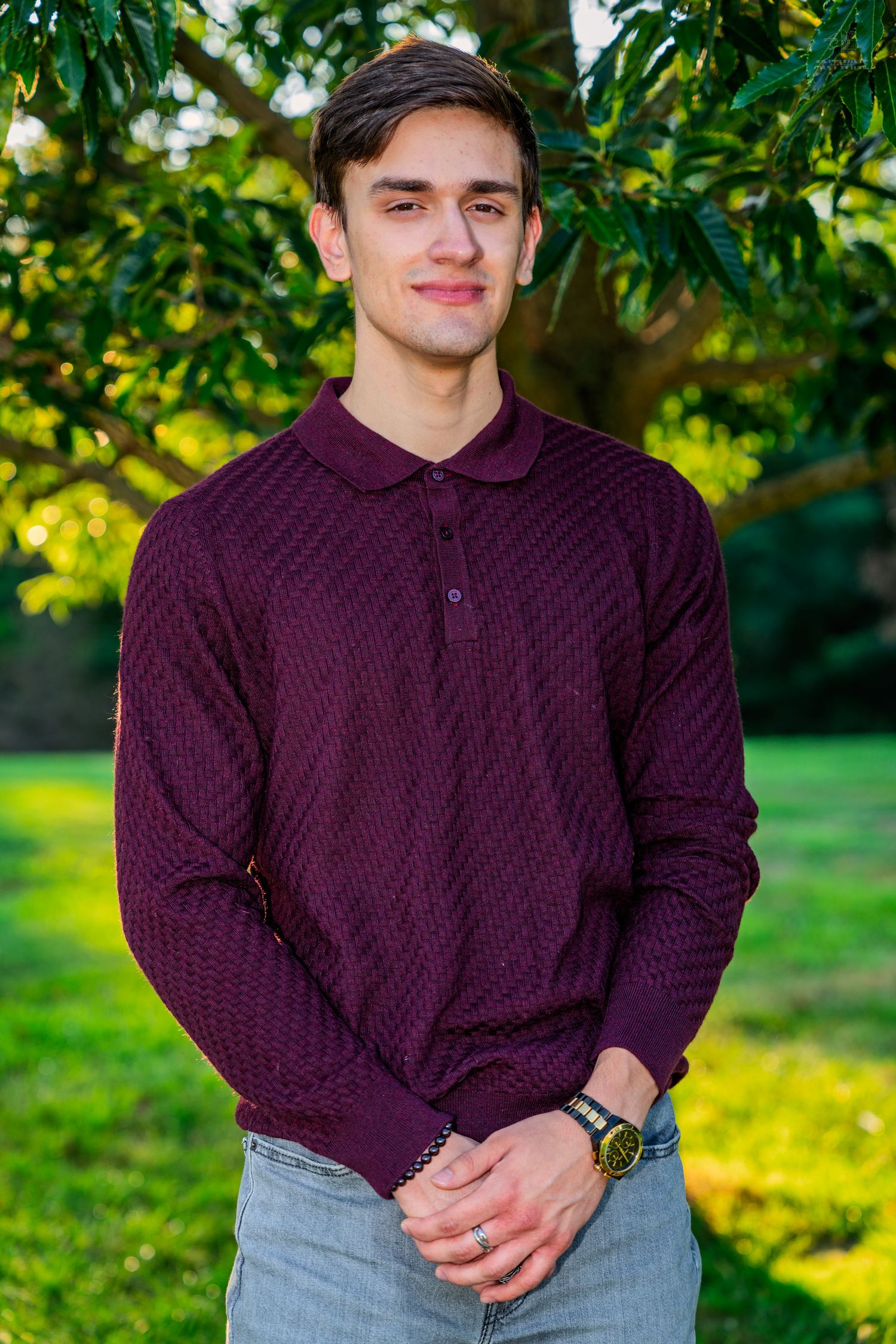 Young man in burgundy polo sweater smiling outdoors with green trees in background.