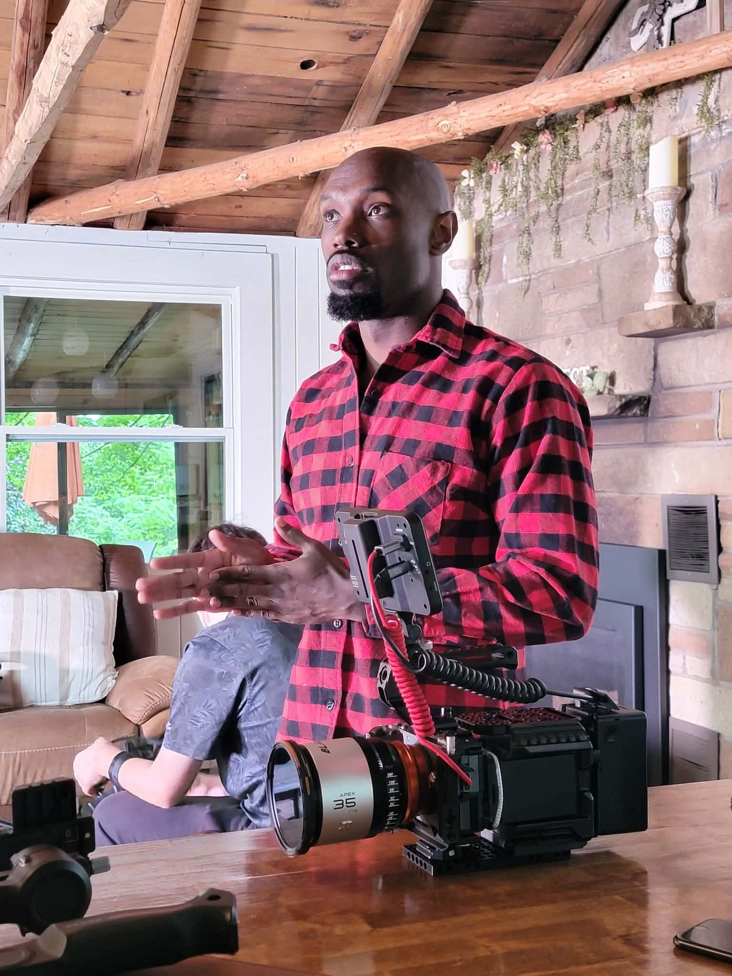 A tall Black man in a red plaid shirt sits behind a professional cinema camera inside a rustic wood-beamed cabin.