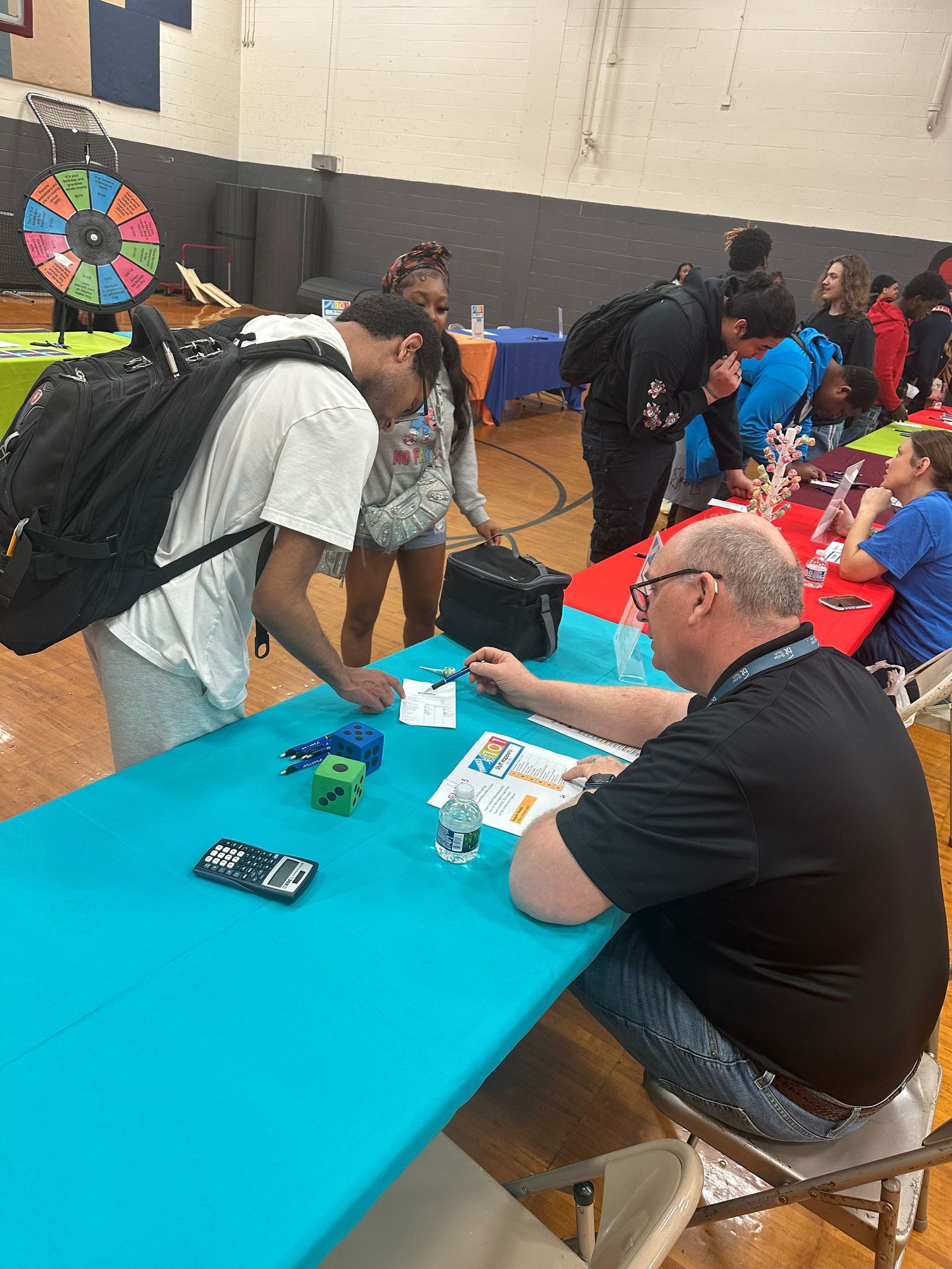 Students interact with a representative at a teal table during a school career fair in a gymnasium.