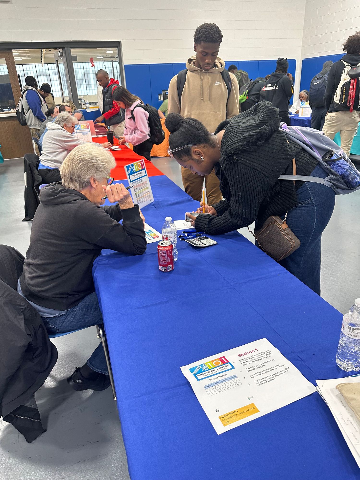 Students and adults gather at blue tables during a school career fair or educational resource event in a gymnasium.