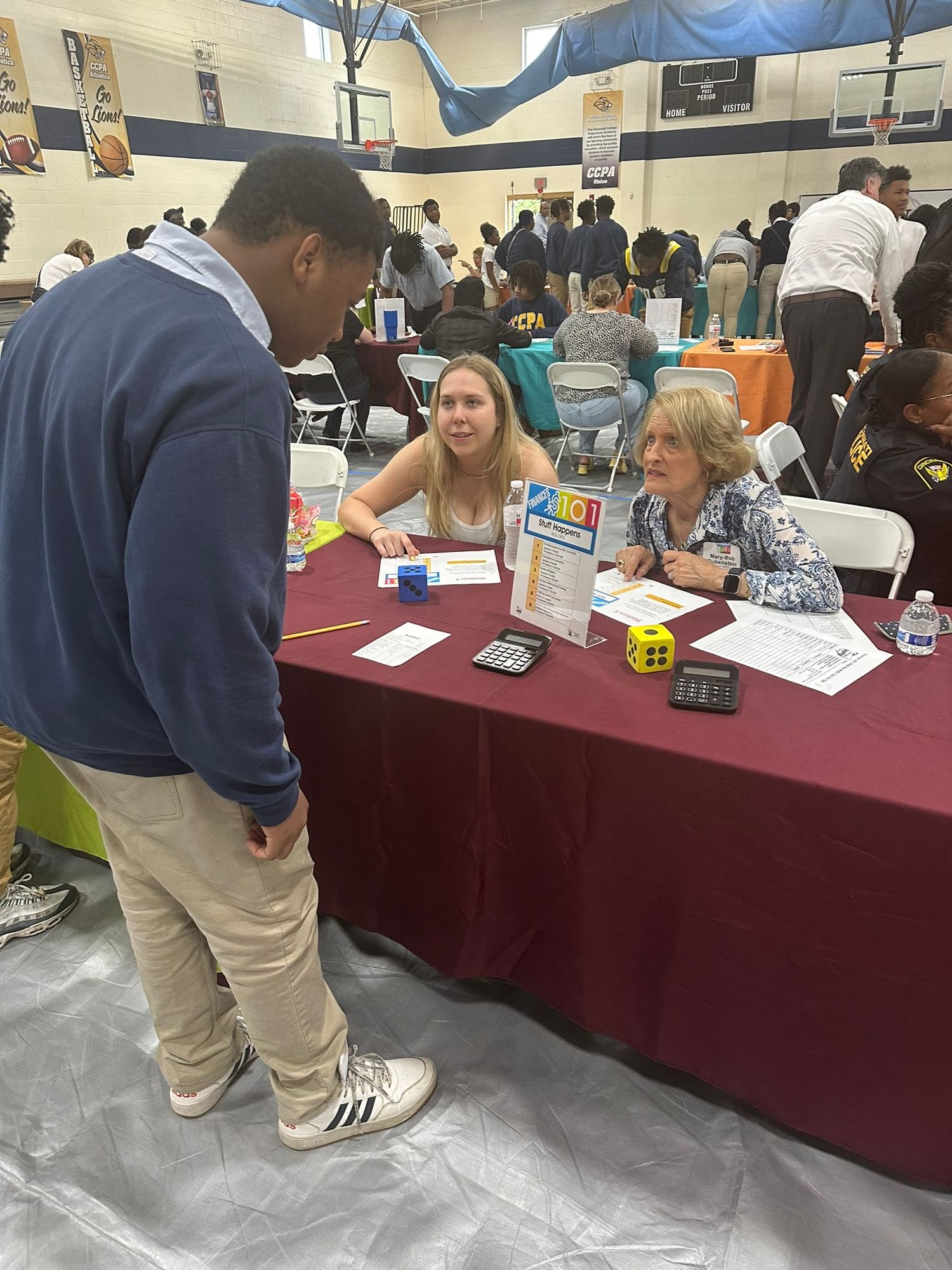 Students and adults engage in a financial literacy fair activity at a table with a burgundy tablecloth in a school gymnasium.
