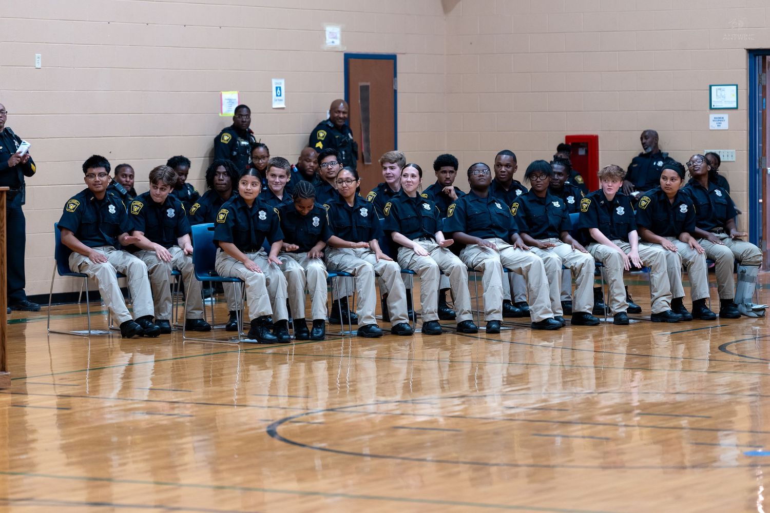 A group of police cadets in uniform sit in rows during a formal ceremony inside a gymnasium.