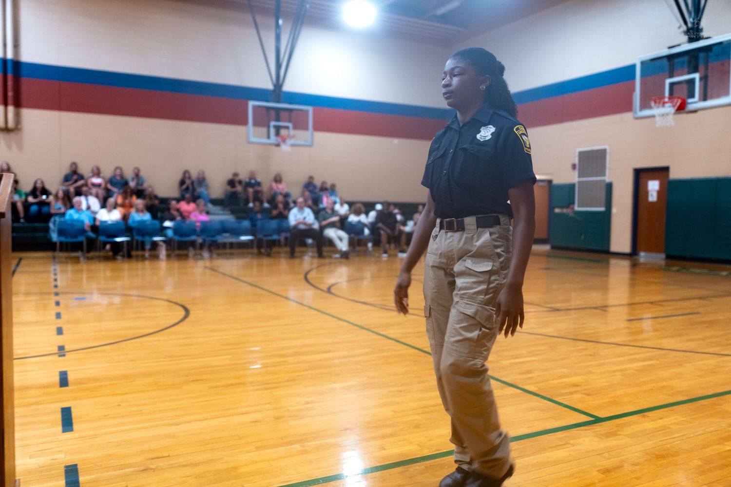 A law enforcement officer stands on a gymnasium floor addressing a seated crowd during a community meeting.