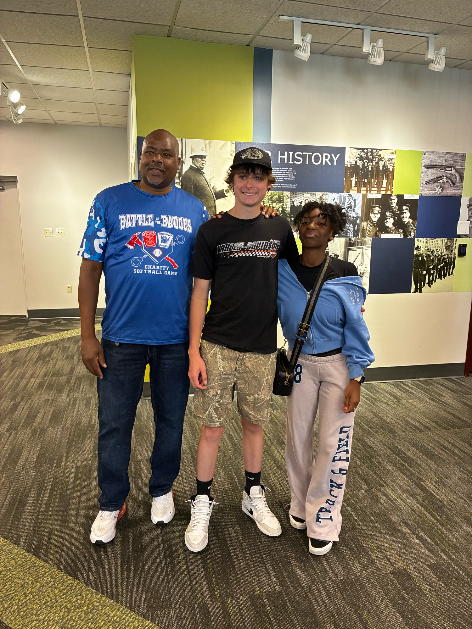 Three people smiling together indoors near a history wall display in what appears to be a museum or educational center.