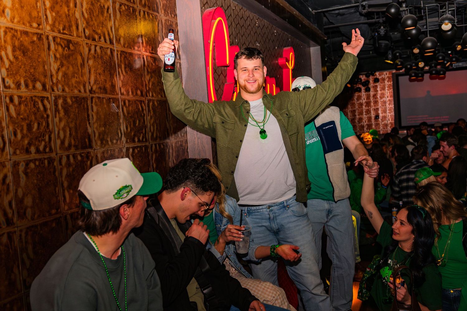 Man in green jacket celebrates St. Patrick's Day at a bar with arms raised.