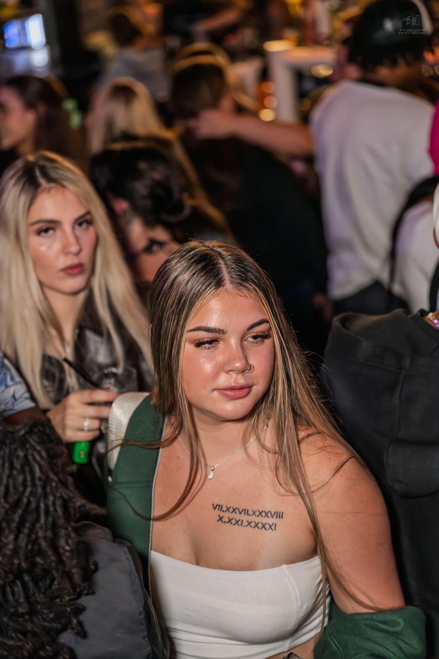 Young blonde woman with a Roman numeral chest tattoo at a crowded nightlife event.