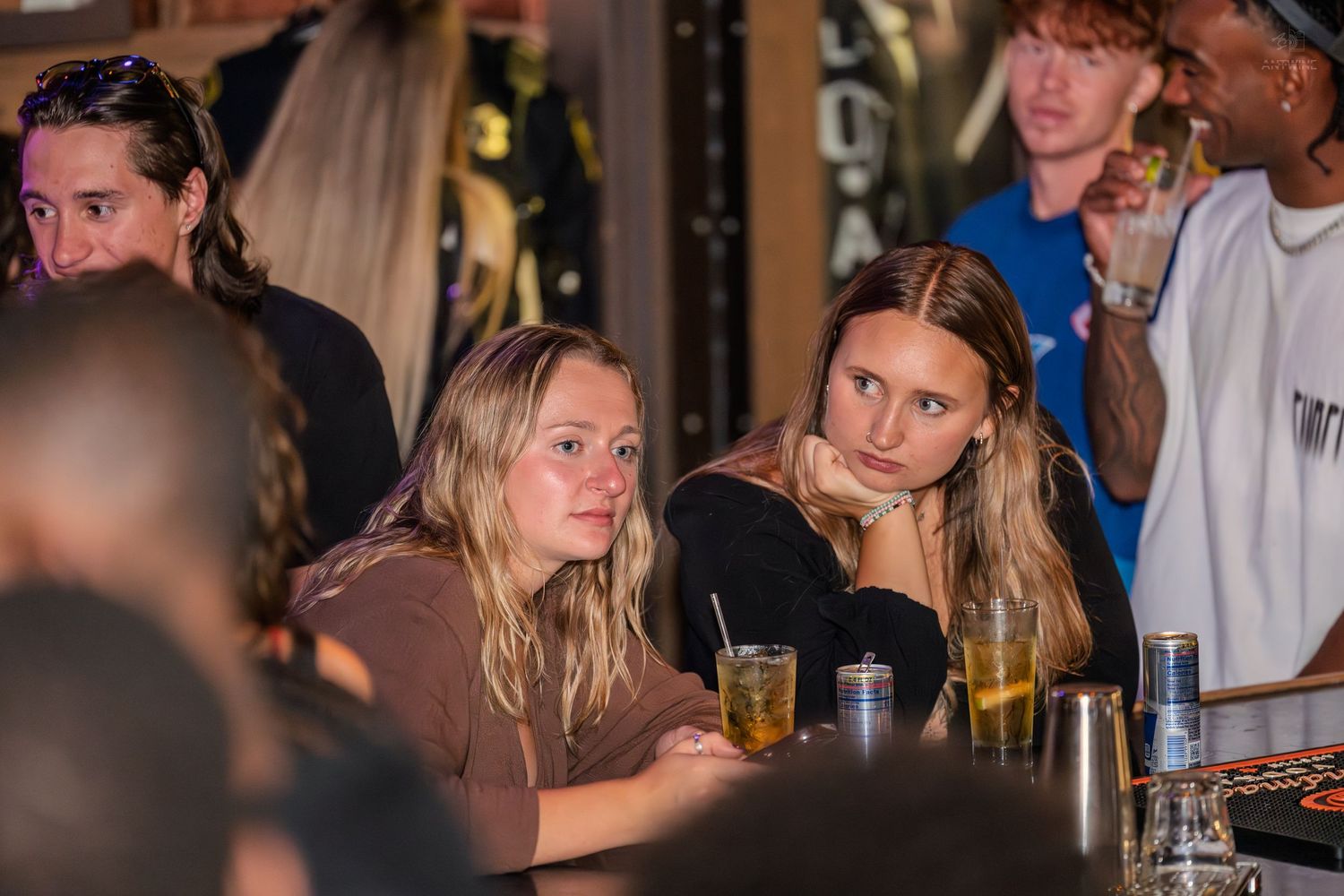 Two blonde women sitting at a bar with drinks at a social event.
