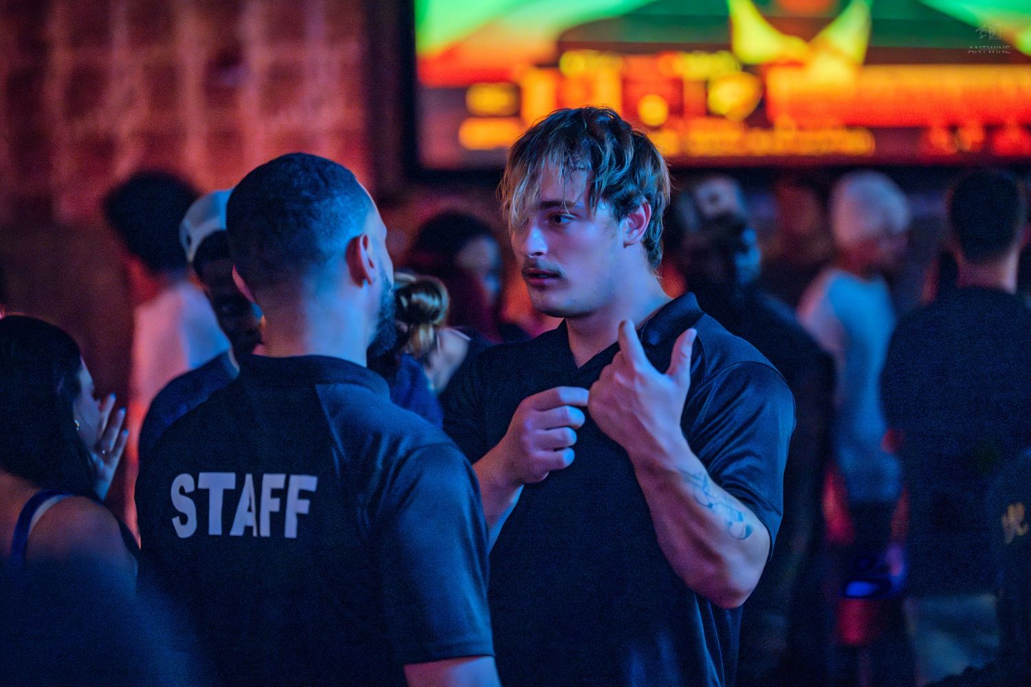 Two men in black shirts talking at a nightclub, one wearing Staff uniform.